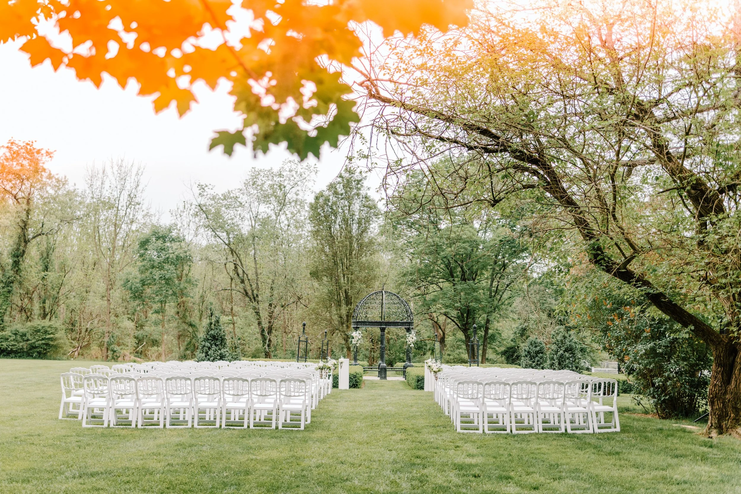 Outdoor wedding setup with rows of white chairs on a green lawn, facing an arch decorated with white flowers, surrounded by trees under a cloudy sky.
