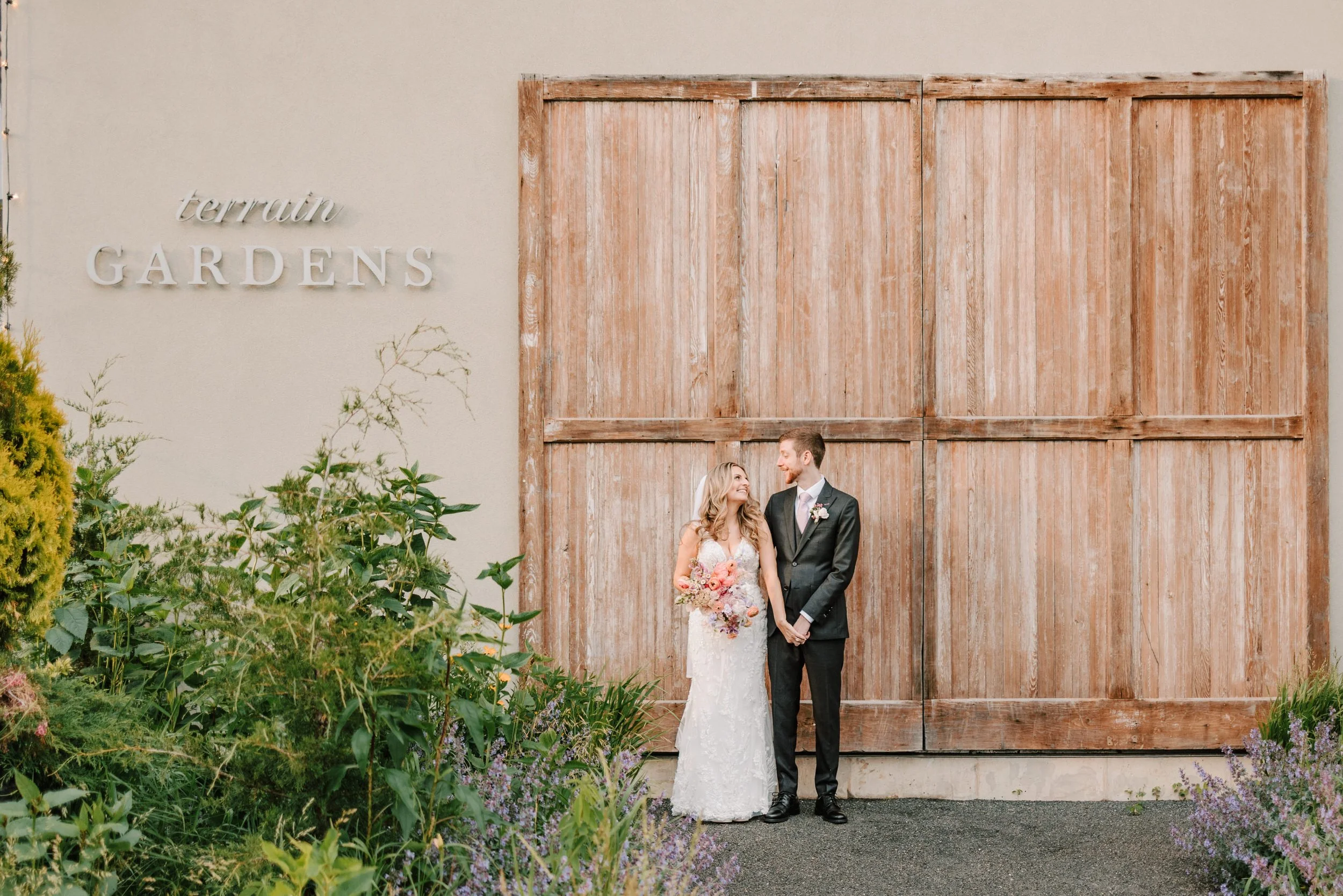 A bride and groom stand side by side in front of a large wooden door at a garden wedding venue, with greenery and purple flowers on the left and right sides. The bride holds a bouquet and looks at the groom, who wears a dark suit and looks back at her. The venue sign reads "terrain GARDENS."