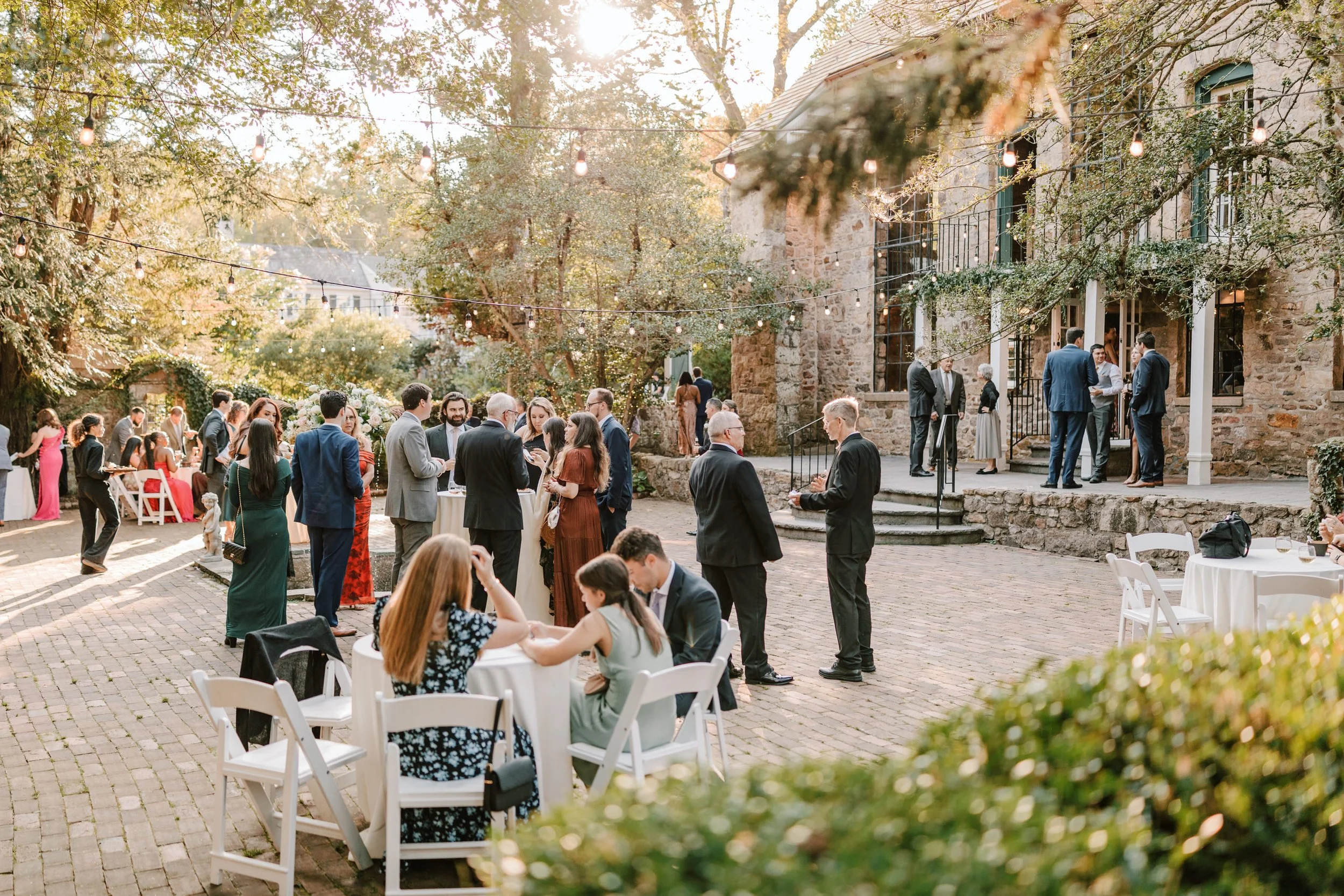 Guests attending an outdoor wedding reception in the courtyard of a rustic stone building, with string lights hanging above, tables and chairs set up for mingling and dining, surrounded by lush trees and greenery.