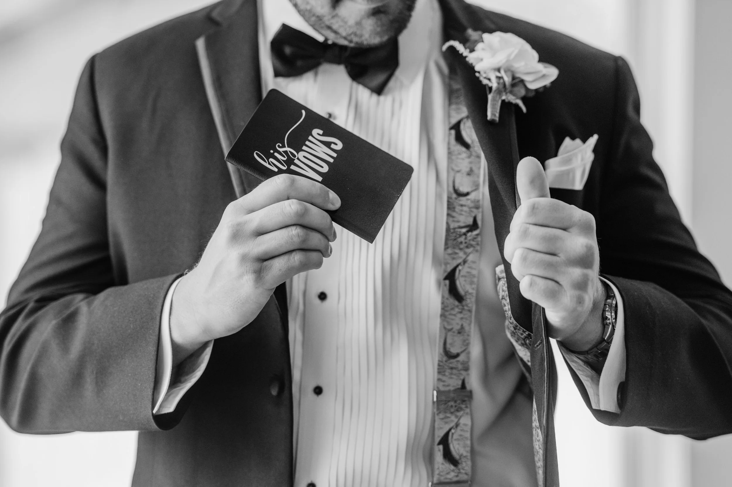 A man in a tuxedo and bow tie holding a card that says "his vows" in one hand, giving a thumbs up with the other.