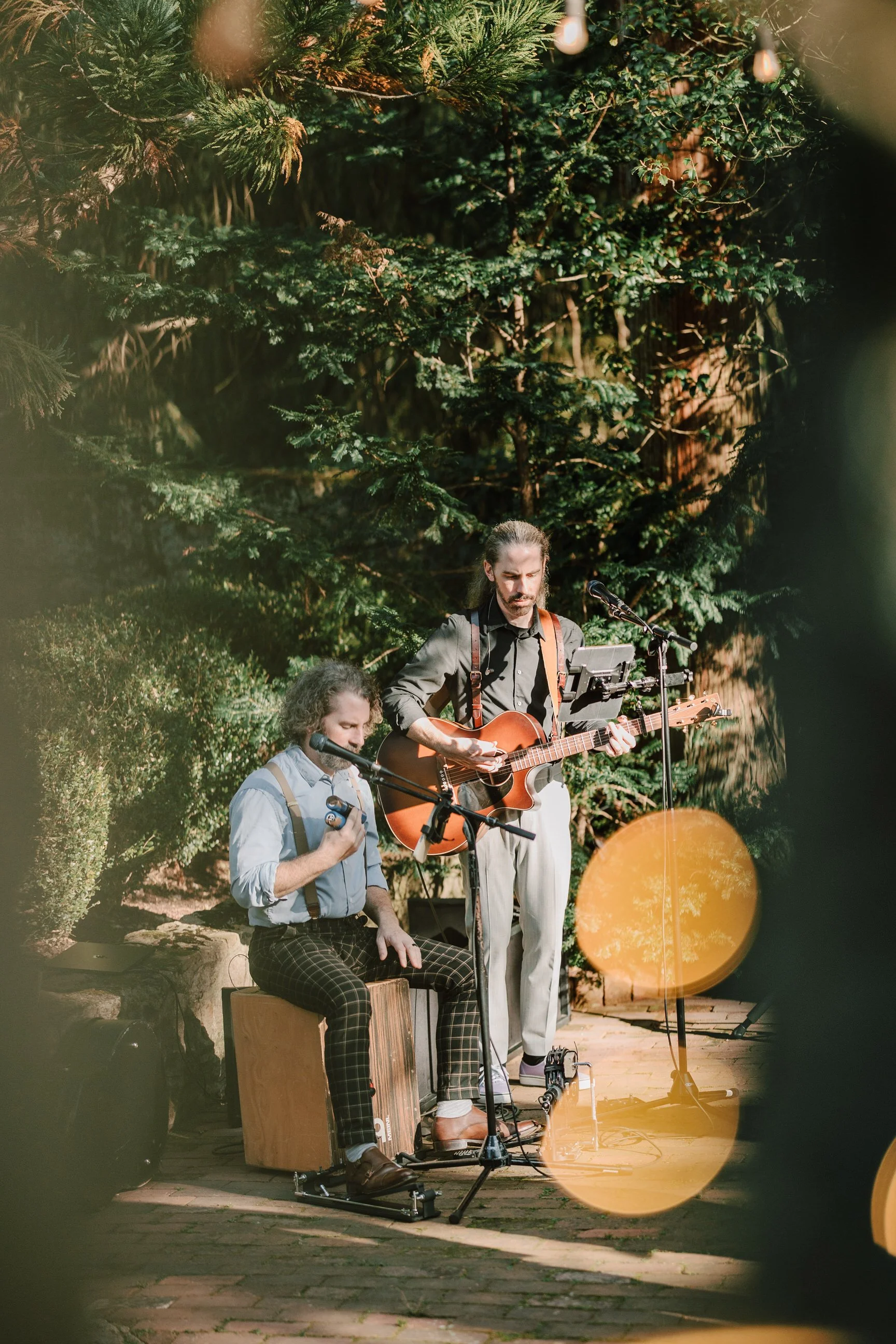 Two musicians performing outdoors, one sitting on a cajón and the other standing, playing guitar, with greenery and trees in the background and decorative string lights overhead.