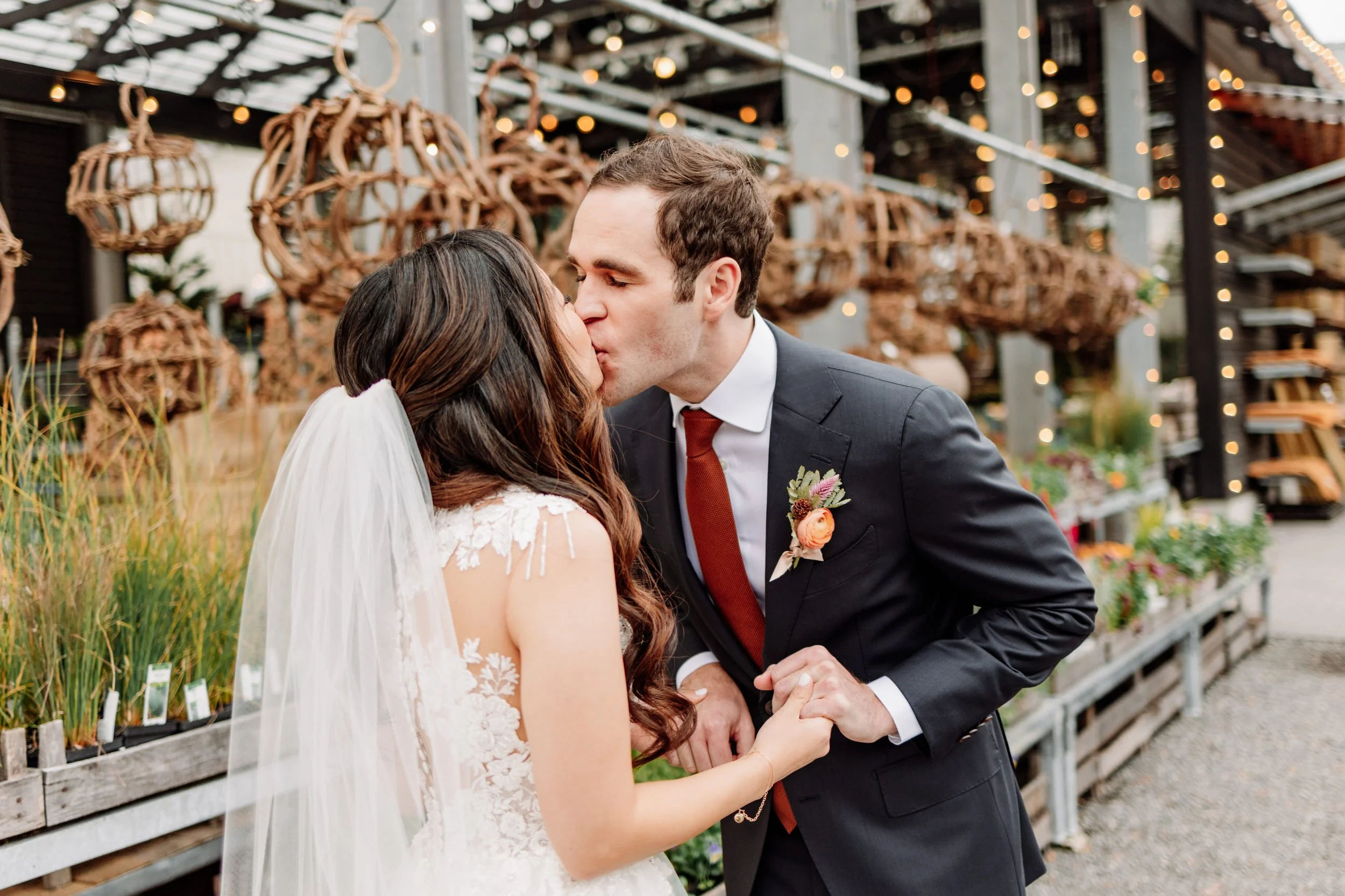 A bride and groom kiss outdoors at their wedding, holding hands, with decorative hanging lights and plants in the background.
