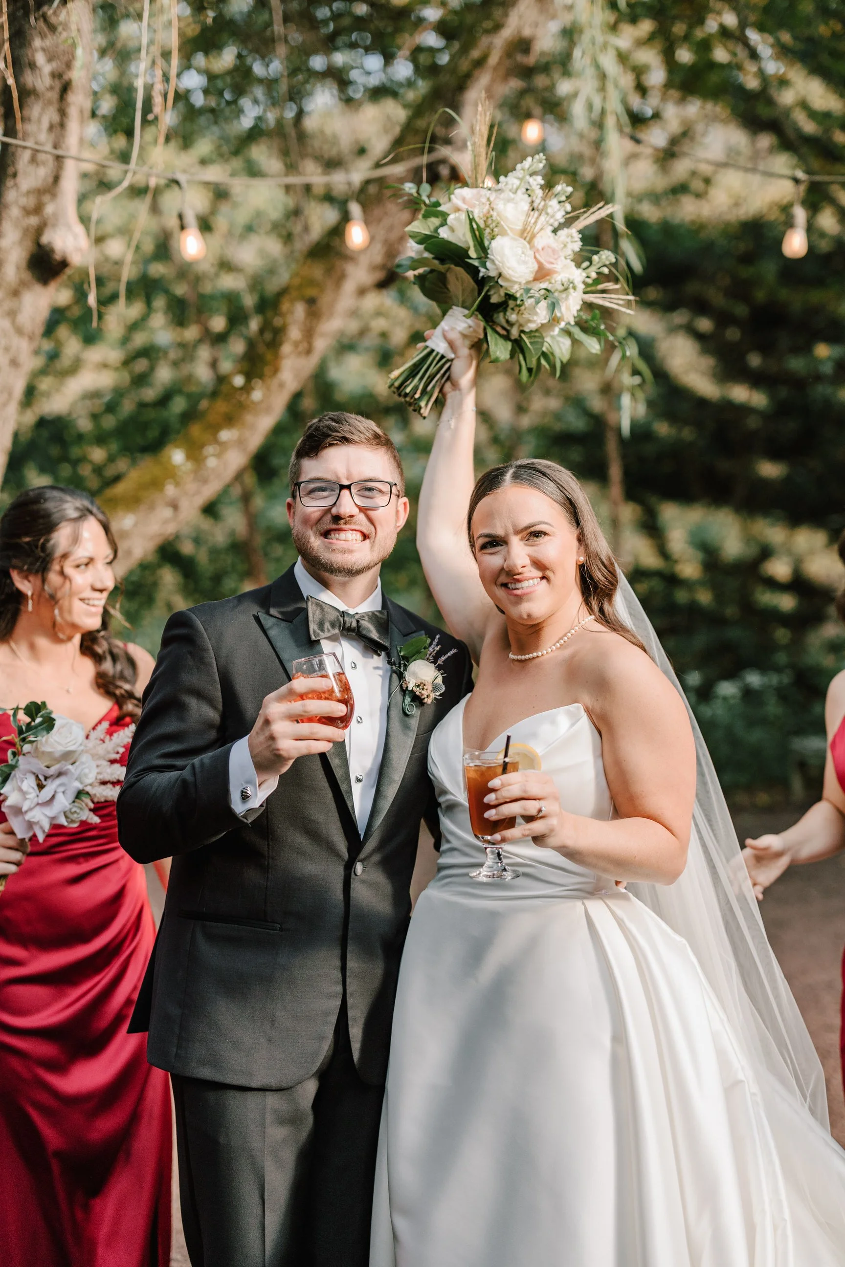 A bride and groom celebrate at their wedding outdoors, with the bride holding a bouquet high above her head, both holding drinks, and smiling. Bridesmaids are in the background.