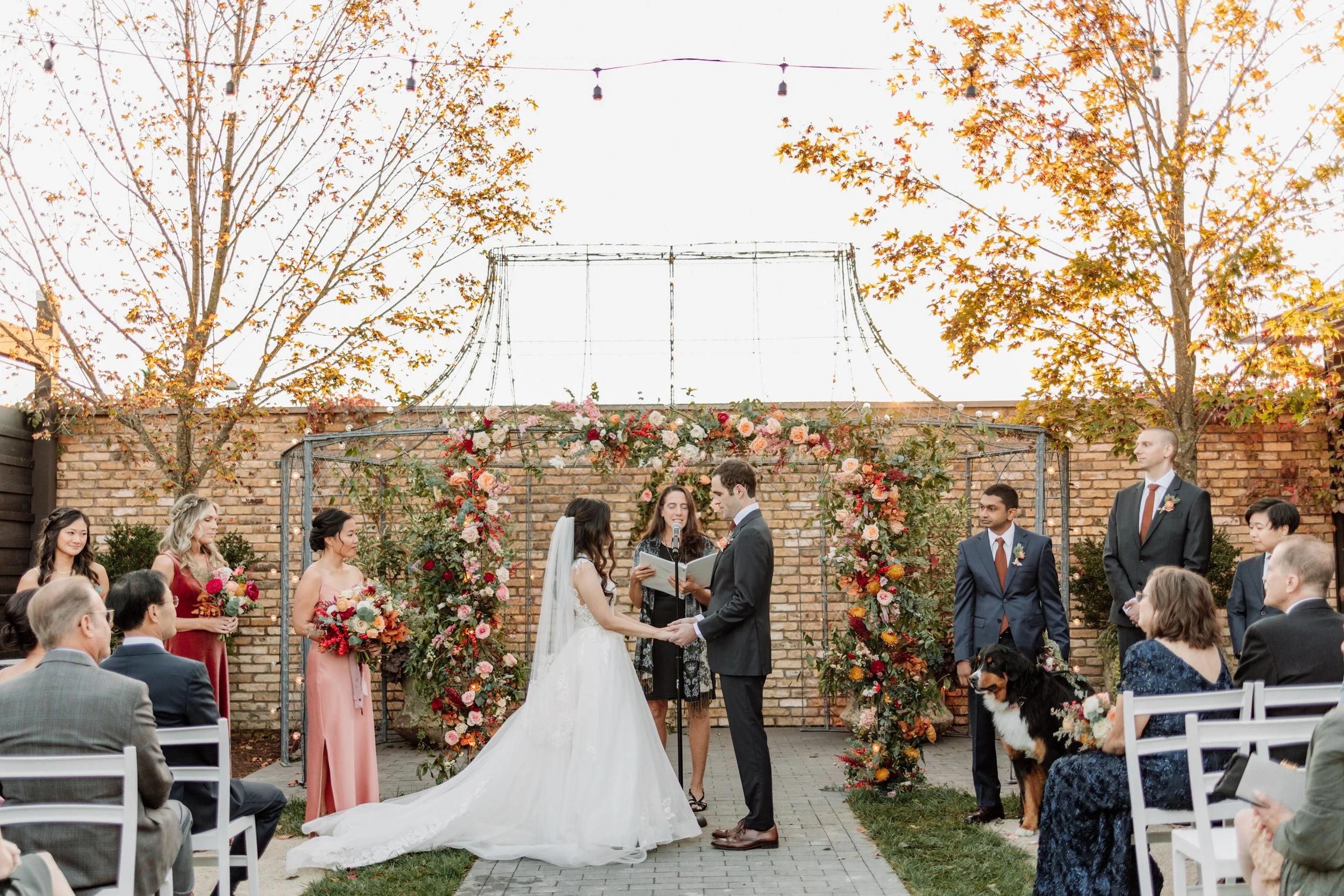 A wedding ceremony taking place outdoors with the bride and groom holding hands, surrounded by bridesmaids and groomsmen. There is floral decoration and a brick wall backdrop, with guests seated on either side.