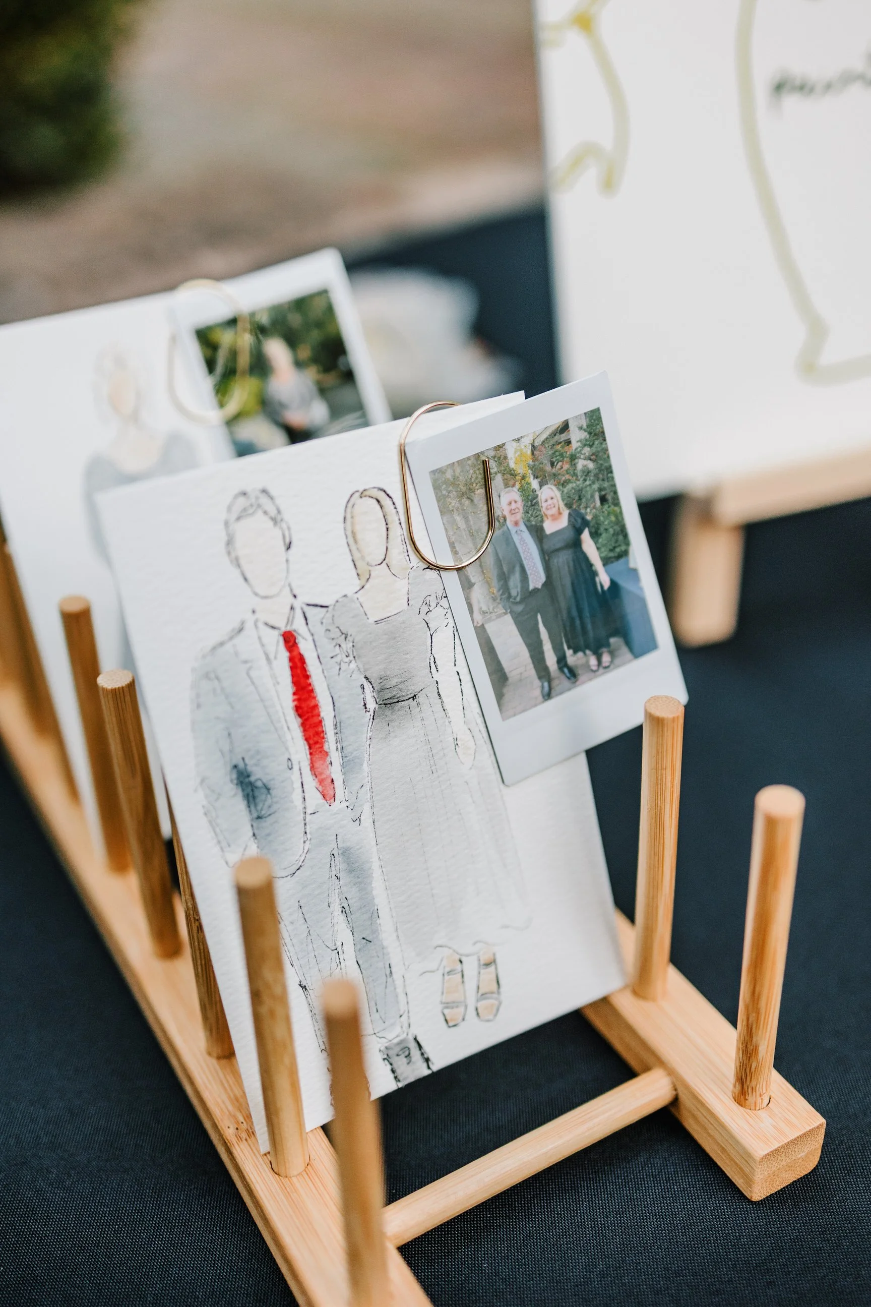 A wooden photo display stand holding a sketch of a couple and two printed photos, one clipped with a paperclip. The photos feature people outdoors.