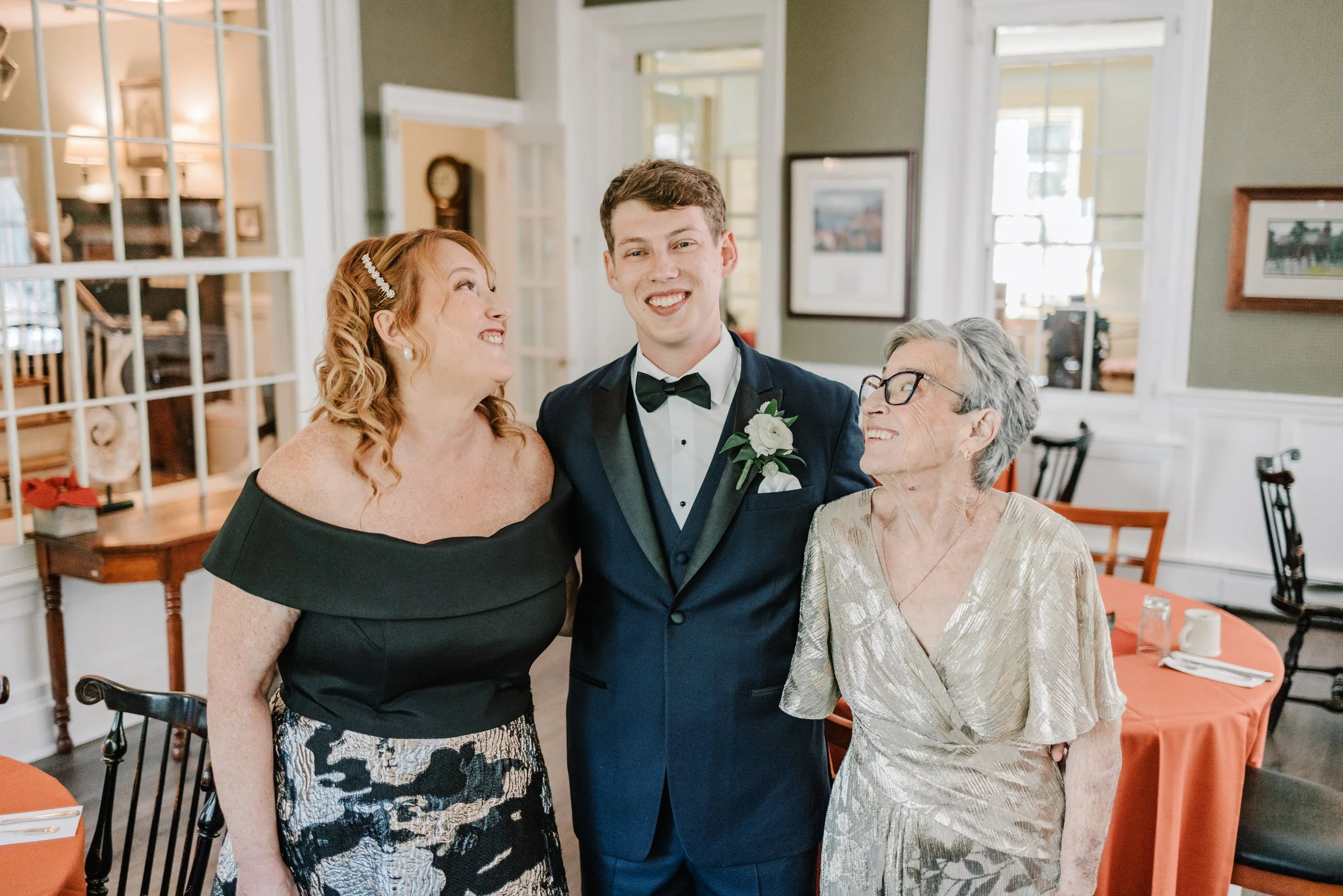A young groom in a tuxedo is smiling with two women, one on each side, at a wedding reception in a decorated venue.