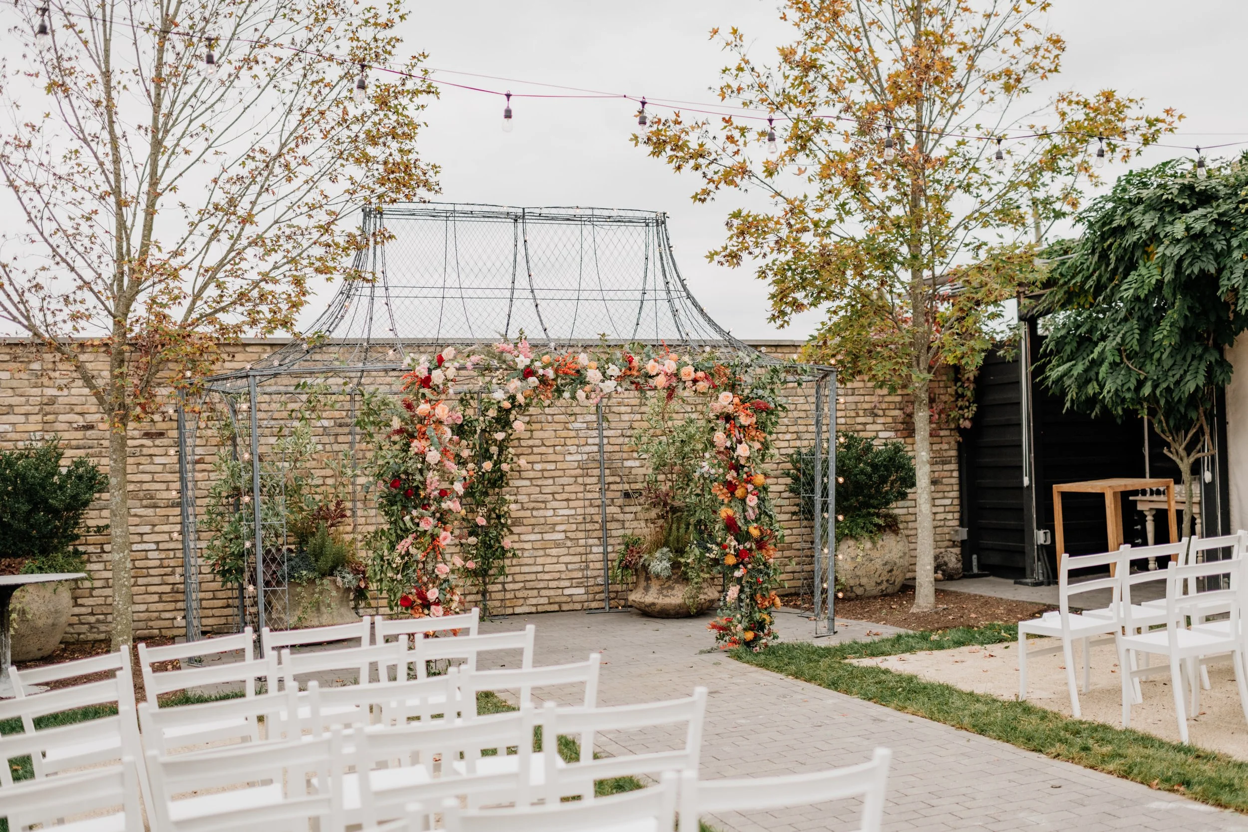 Outdoor wedding ceremony setup with white chairs arranged in rows, a floral arch with pink, white, and red flowers, string lights hanging above, trees with fall foliage, and a brick wall in the background.