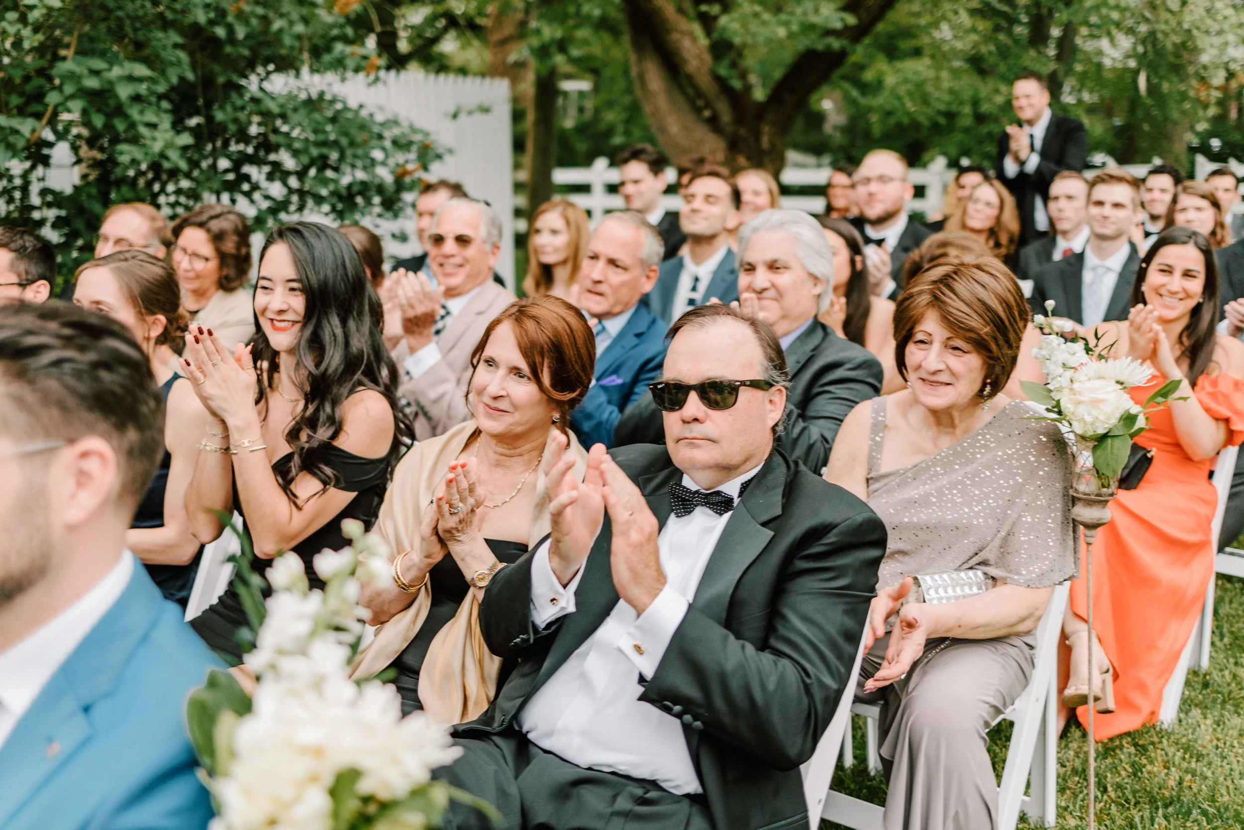 Group of people seated outdoors at a wedding or formal event, clapping and smiling, with some holding bouquets of flowers, greenery and trees in the background.