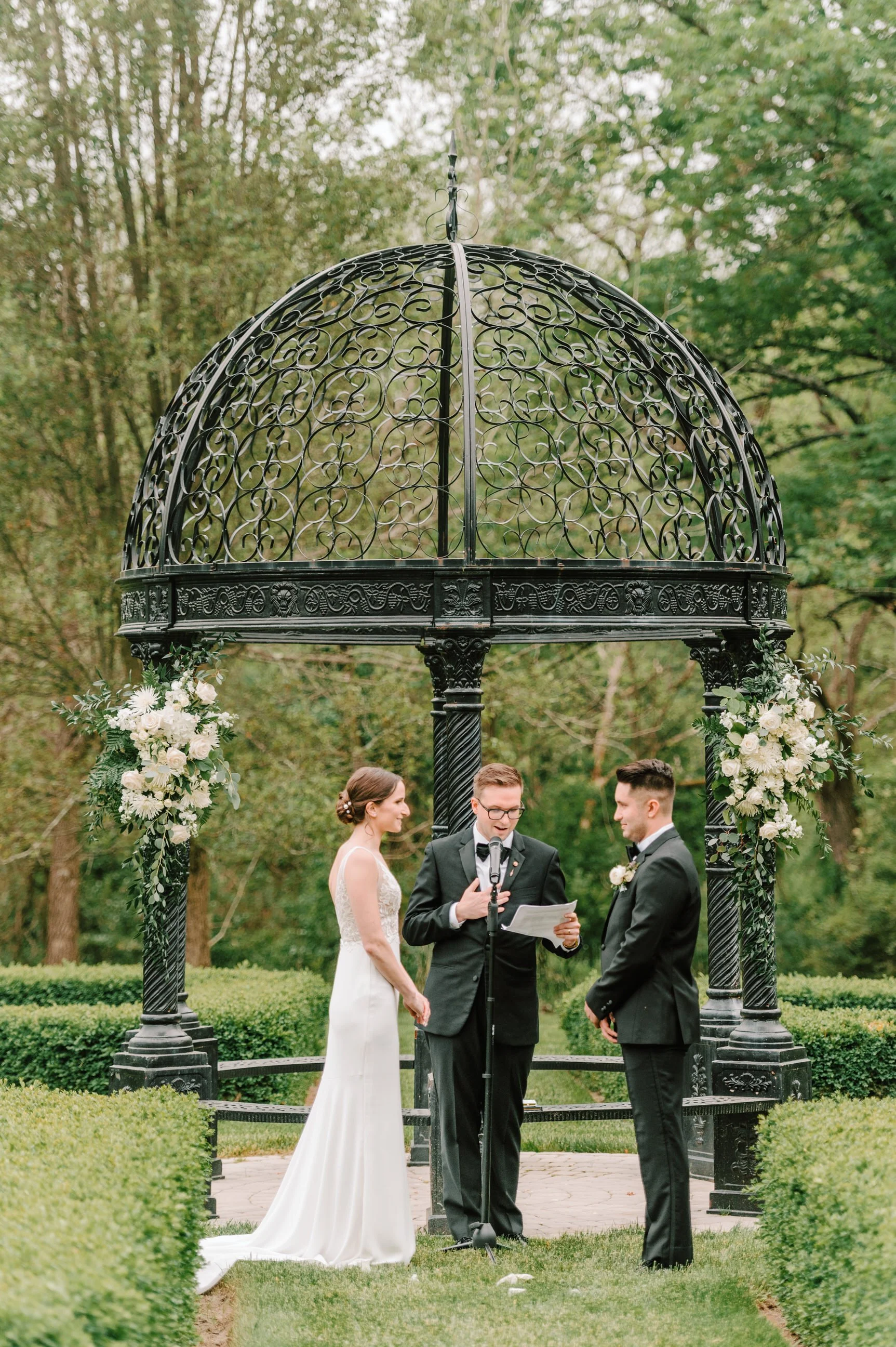 A wedding ceremony taking place outdoors under a decorative black metal gazebo with floral arrangements, with a bride, groom, and officiant.