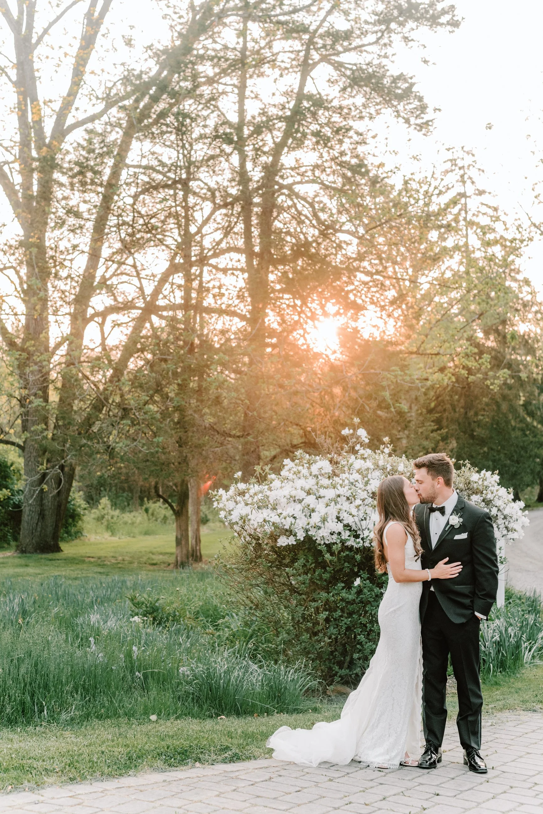 Bride and groom share a kiss outdoors at sunset, surrounded by greenery and blooming flowers.