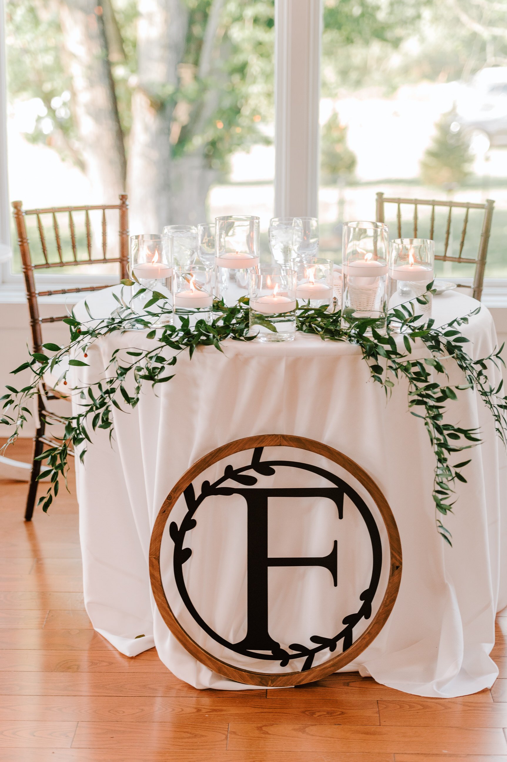 Wedding or event table with a white tablecloth, green leafy garland, glass candle holders with floating candles, and a decorative circular sign with the letter 'F' inside a wreath design leaning against the table, with chairs and large windows in the