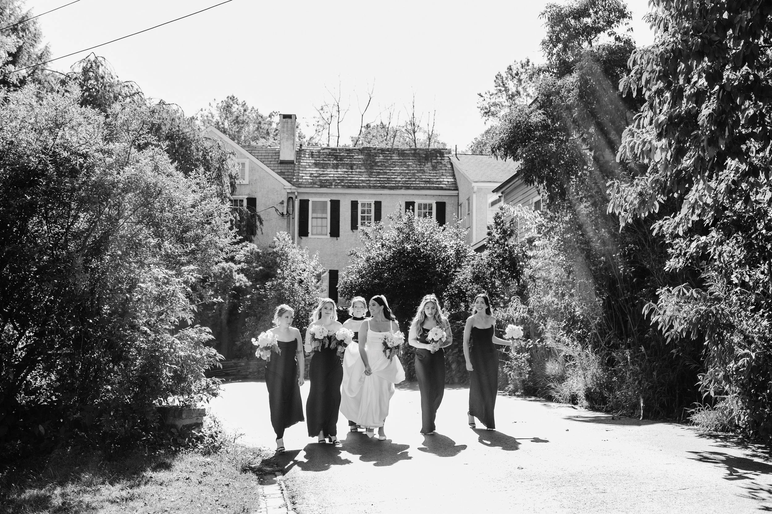 A group of women, including a bride in a white wedding dress, walking down a street with trees and houses in the background, sunlight streaming through the trees.
