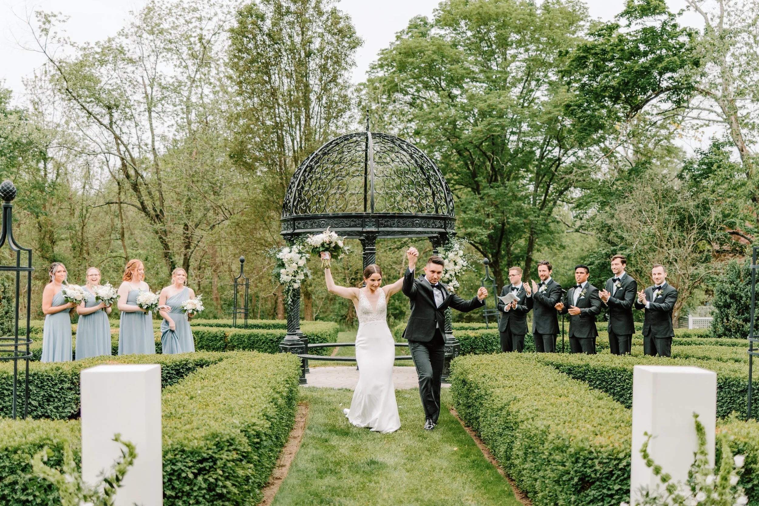A newlywed couple walking down the aisle, holding hands and smiling, sunny outdoor wedding with bridal party and lush green garden.