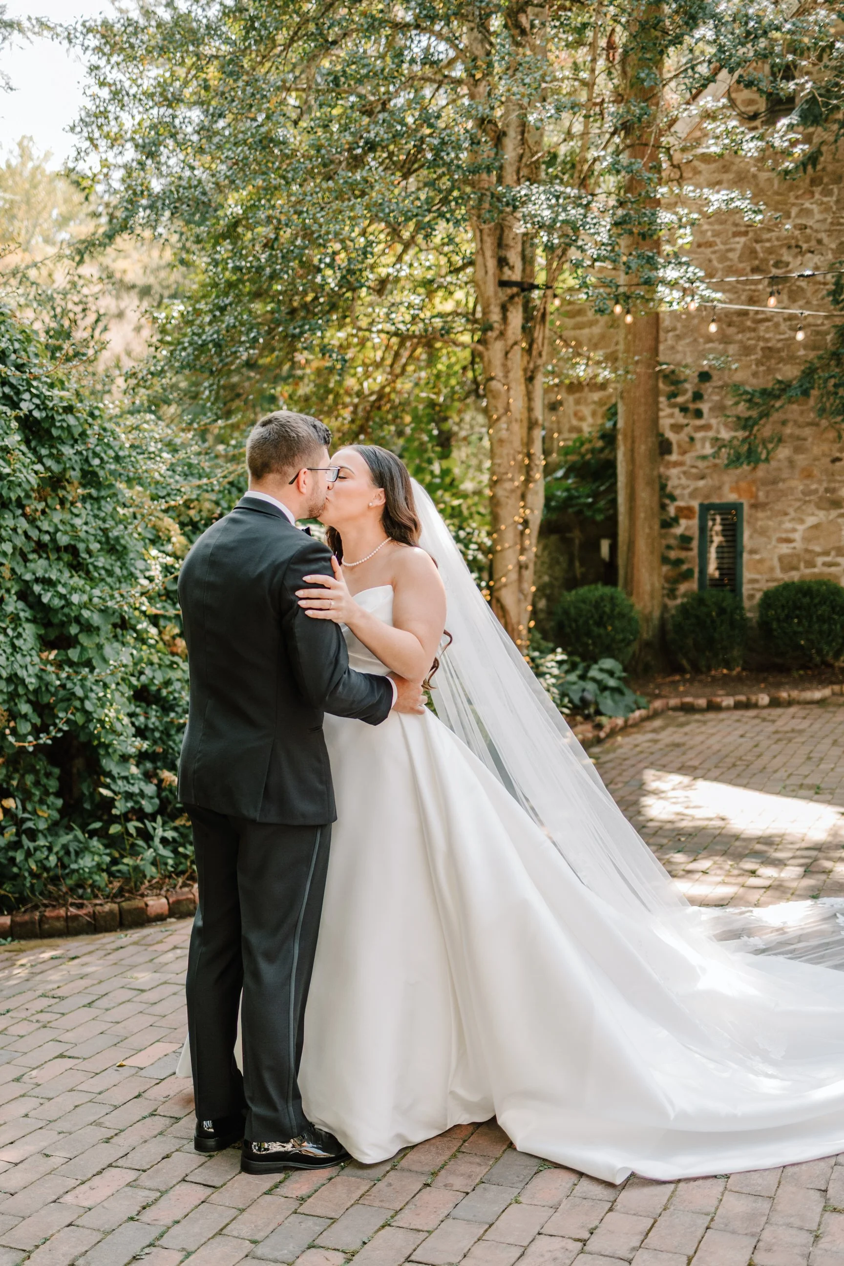 A bride and groom share a kiss outdoors during their wedding, surrounded by greenery and warm lighting.