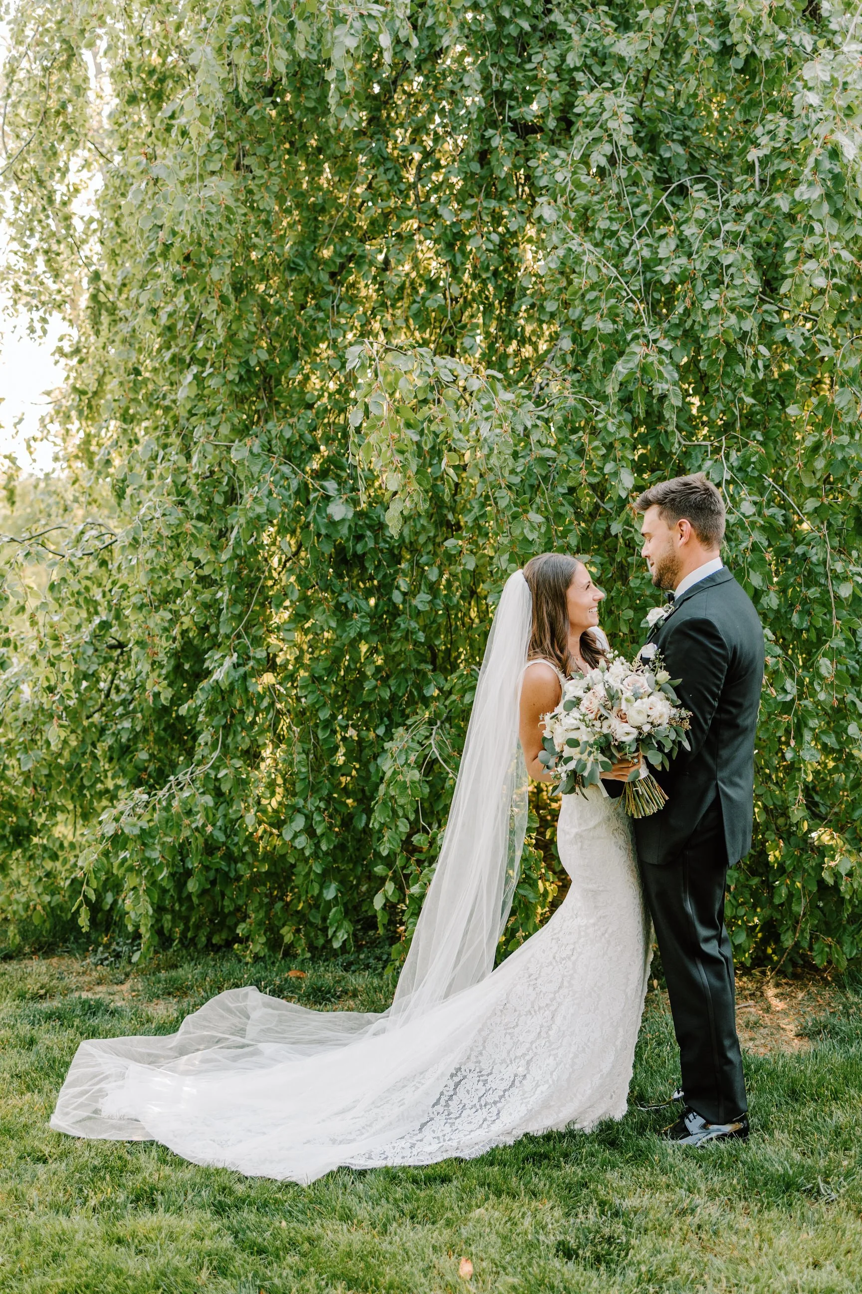 Bride and groom standing on grass in front of a large green leafy bush, smiling at each other, with the bride holding a bouquet of flowers.