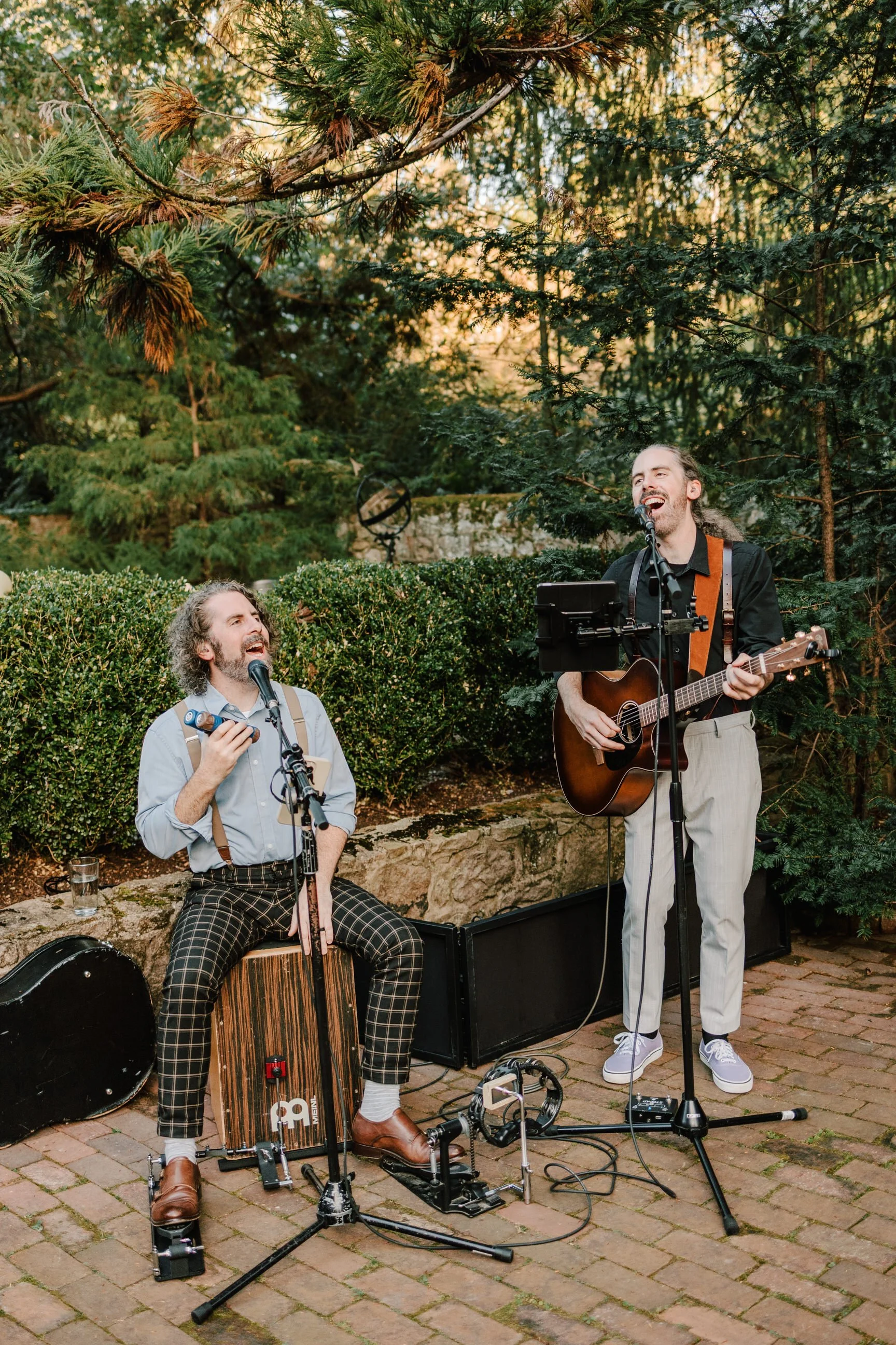 Two male musicians performing outdoors on a brick patio surrounded by lush green trees and bushes, singing into microphones, with one playing an acoustic guitar and the other playing a cajón drum.