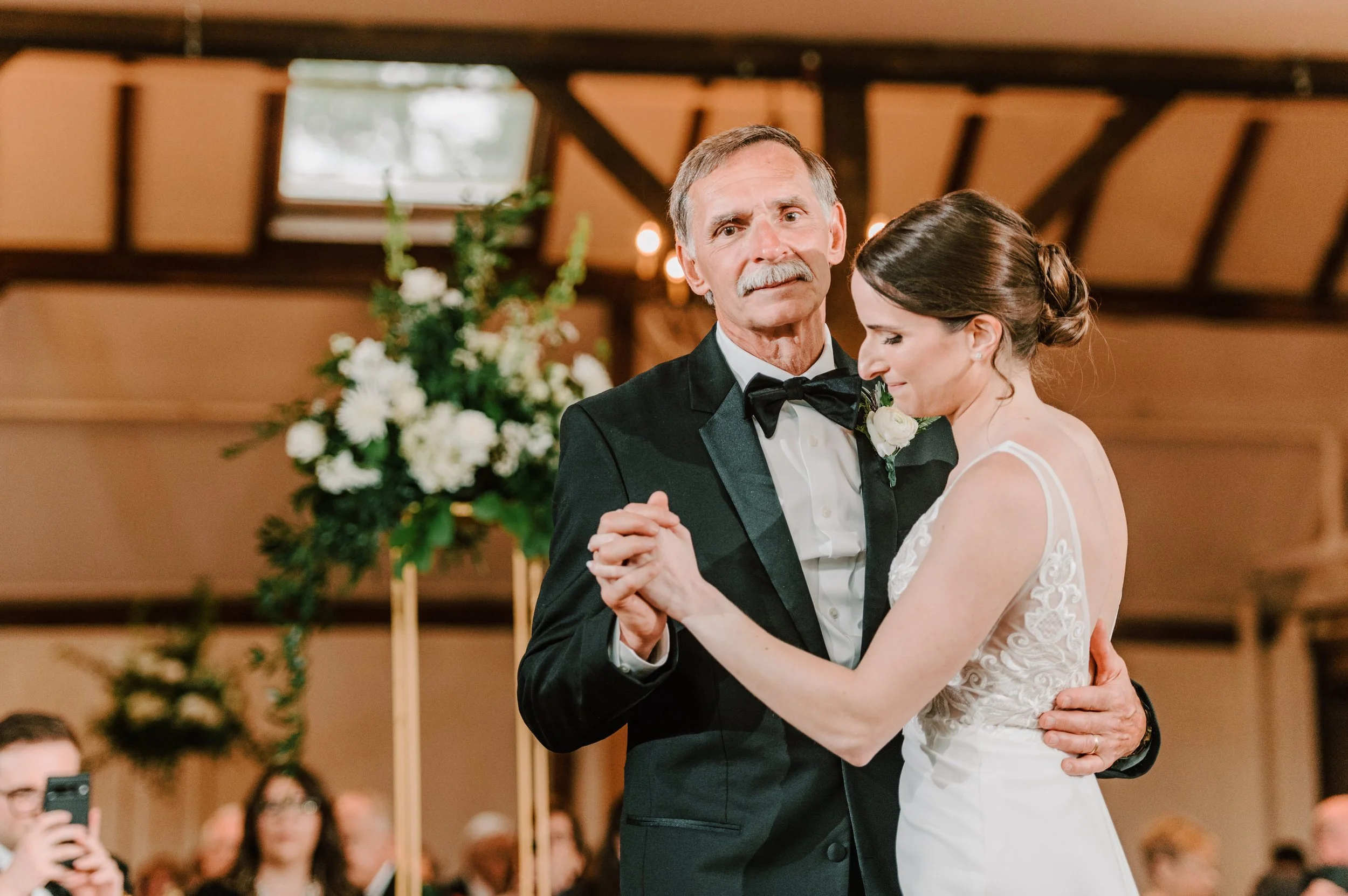 A bride and an older man dance together during a wedding reception. The man is wearing a black tuxedo with a bow tie, and the bride is in a white wedding gown with lace details. They are holding hands and sharing a moment close together, with wedding