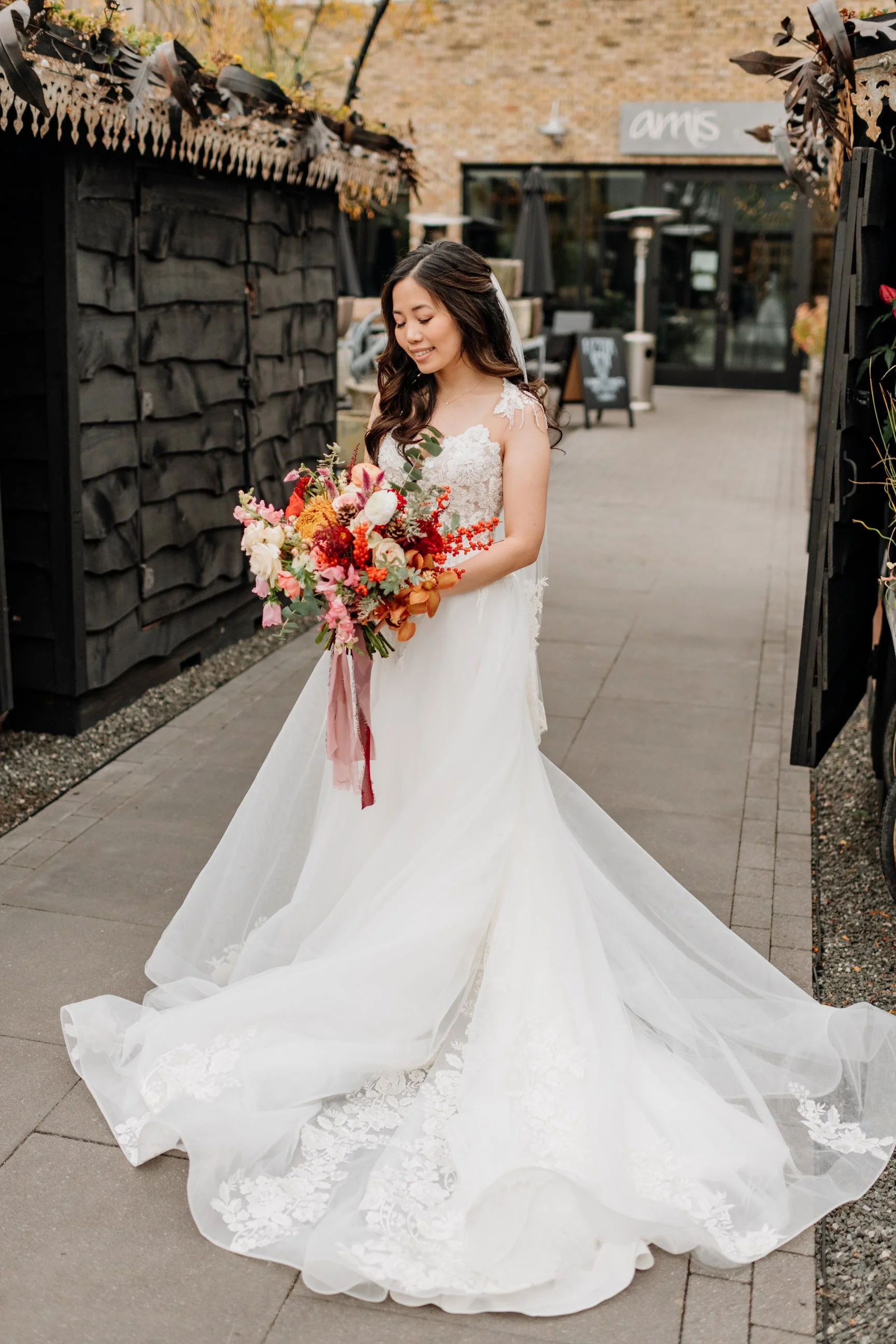 A bride in a white wedding gown holding a colorful bouquet of flowers, standing outdoors on a paved walkway next to black decorative fencing.