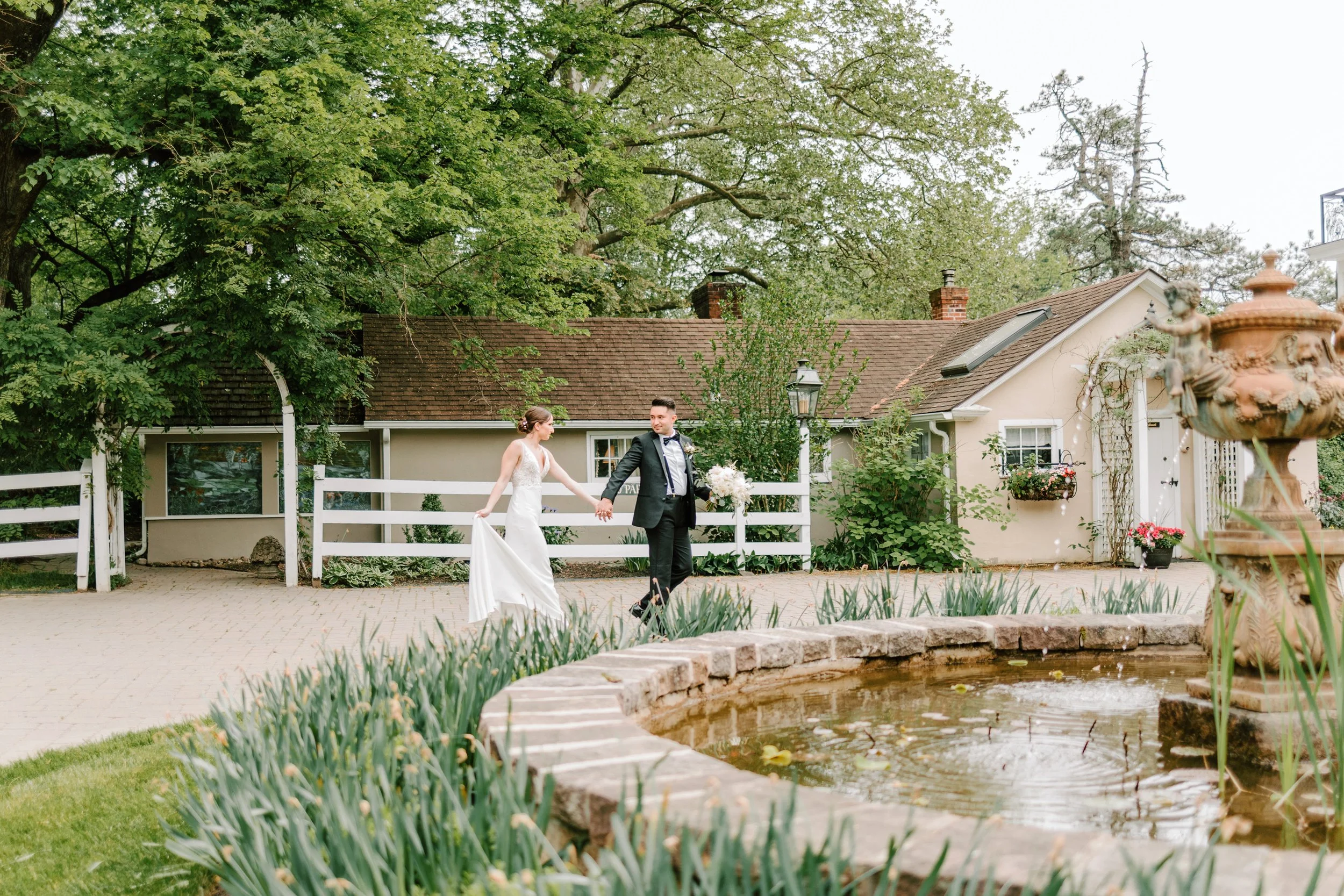 A bride and groom walking hand in hand outdoors in a garden, near a pond with a stone border, greenery, trees, and a small house with potted flowers.