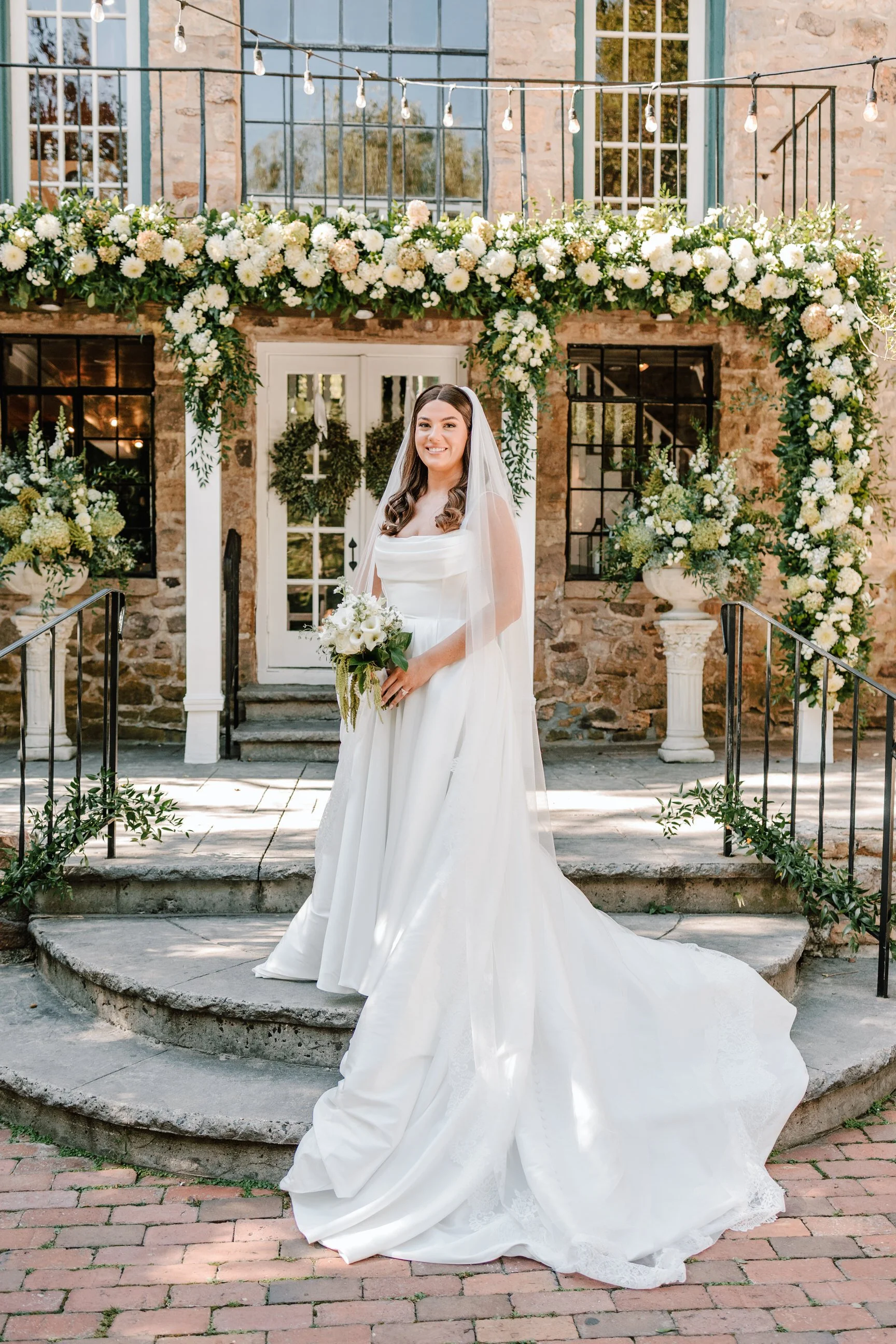 A bride in a white wedding dress holding a bouquet stands on stone steps in front of a floral arch, outside a rustic stone building with large windows, decorated with string lights.