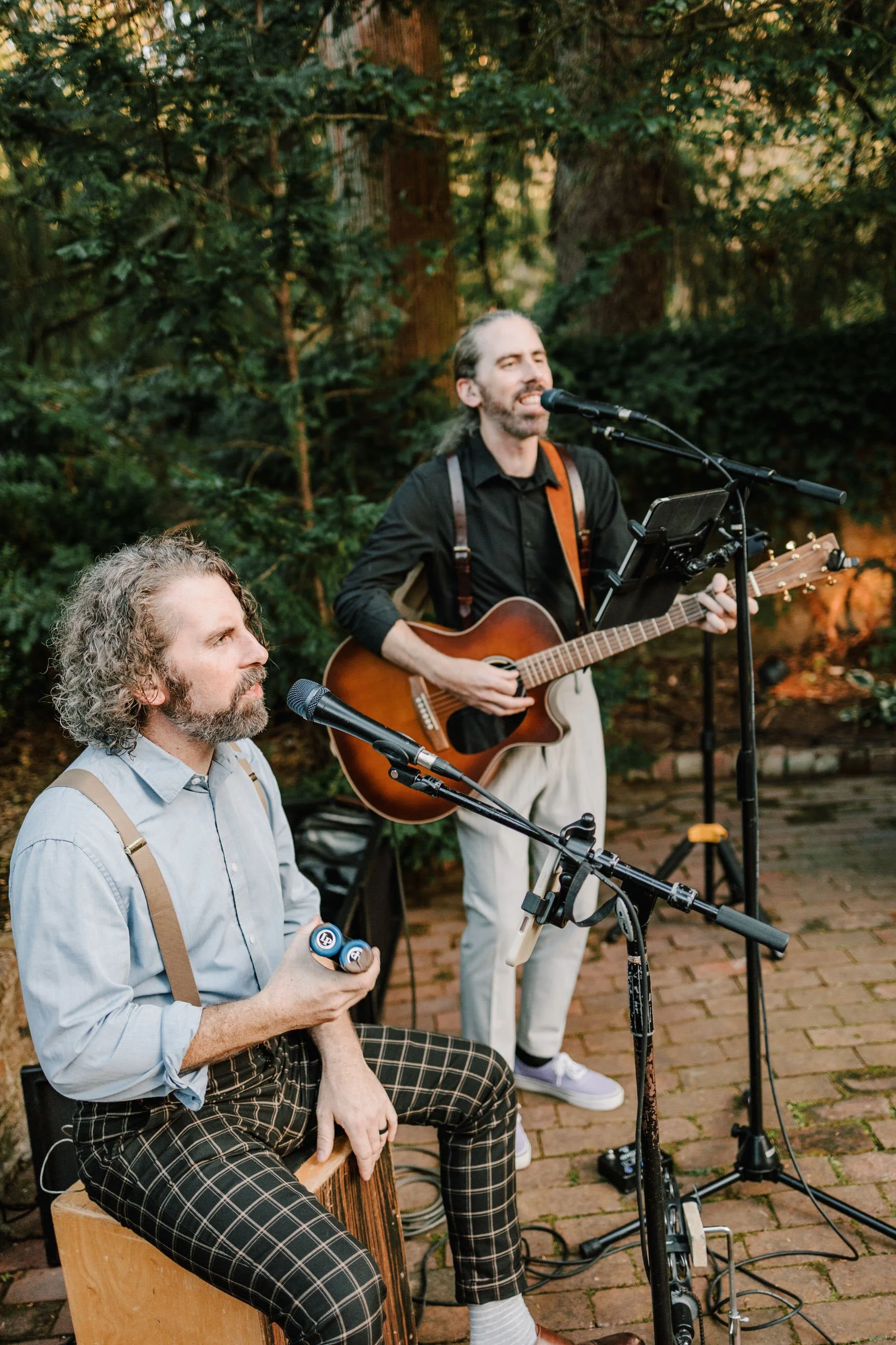 Two musicians performing outdoors; one is seated on a box drum, holding percussion instruments, and the other is standing, singing into a microphone while playing an acoustic guitar, with a lush, green backdrop of trees.