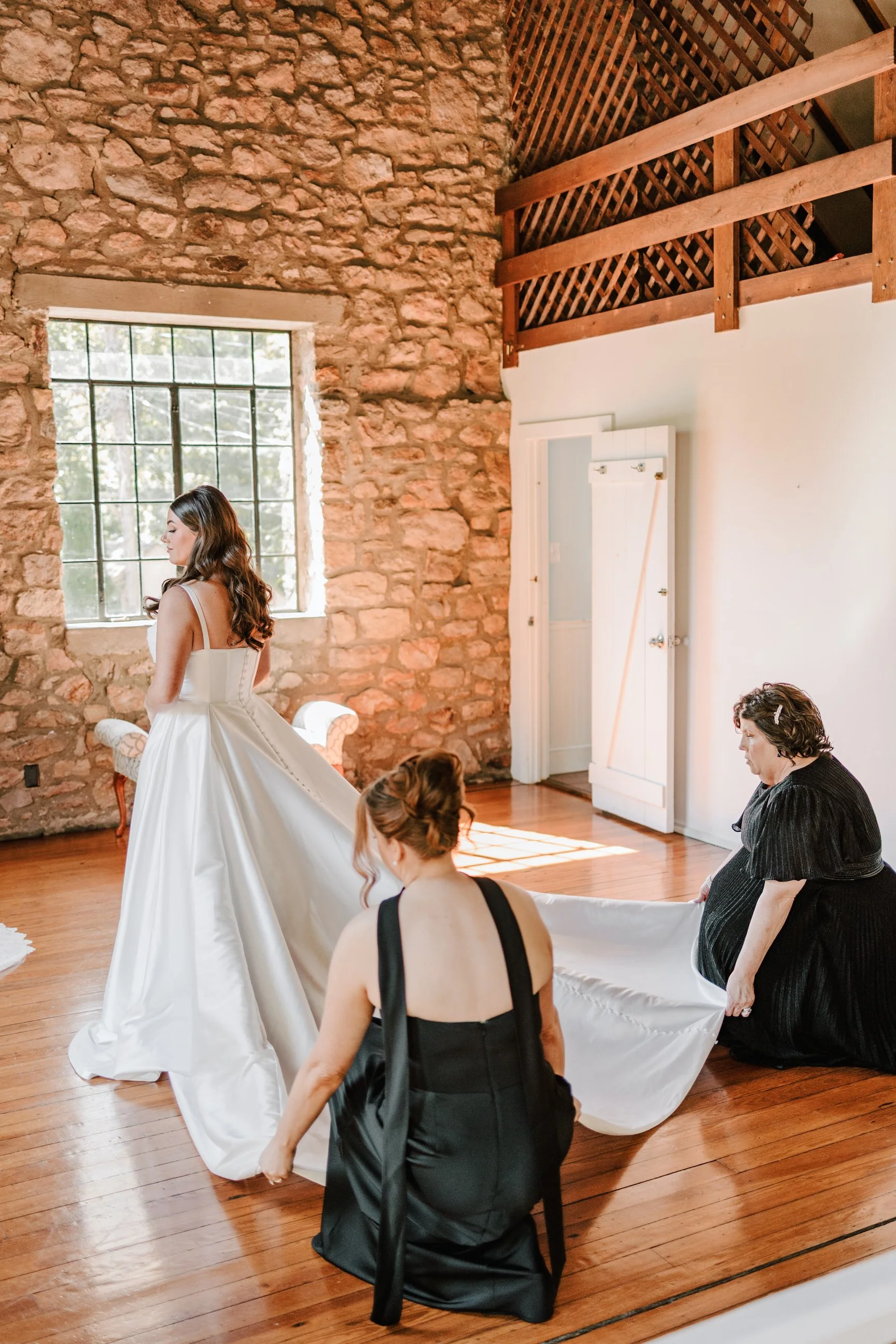 A bride in a white wedding dress is being assisted by two women, one in a black dress and another in a black top, to prepare for her wedding in a rustic room with wooden floors, exposed stone wall, and large window.