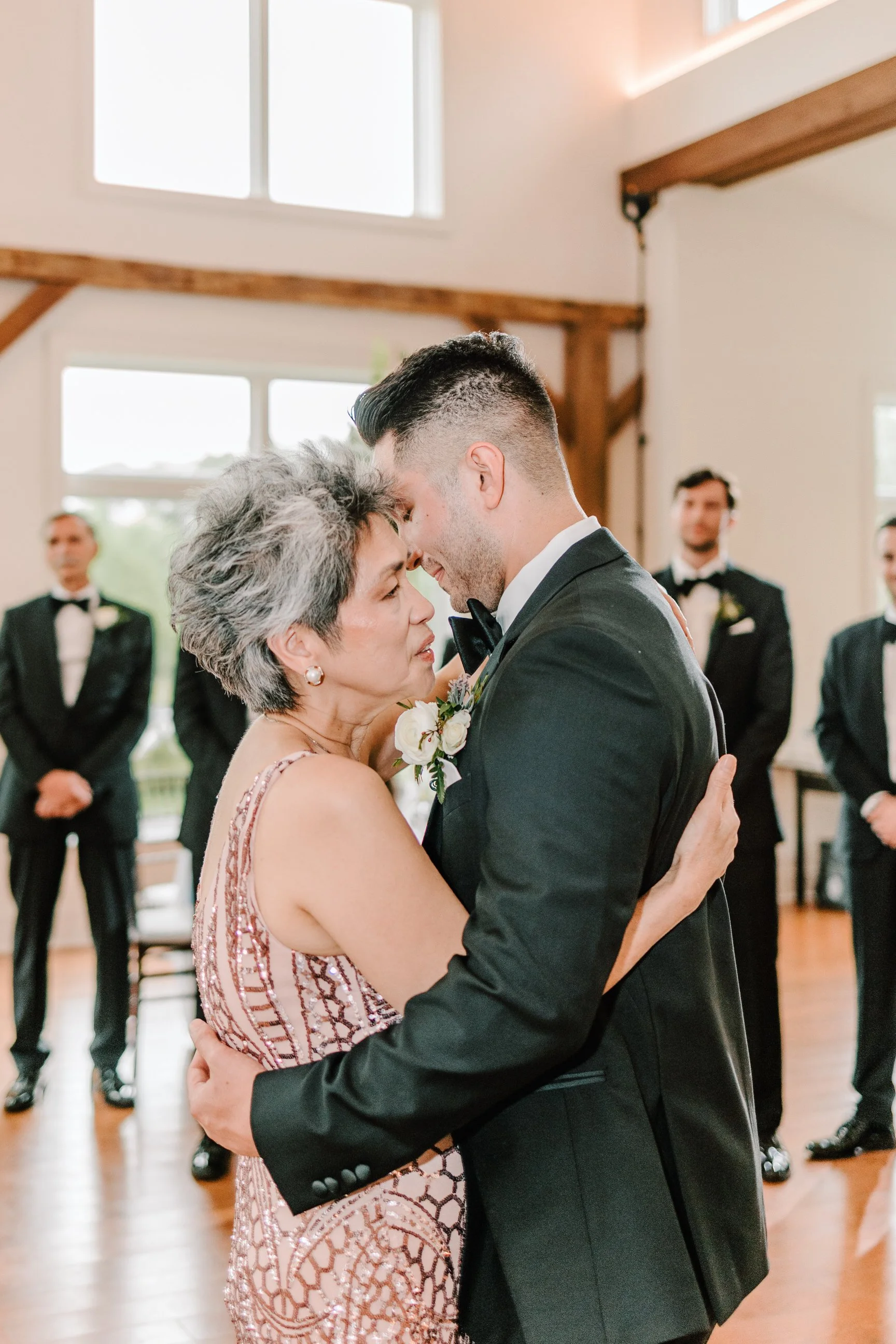 A young man in a tuxedo dances with an older woman in a pink dress during a wedding reception, with other men in tuxedos in the background.