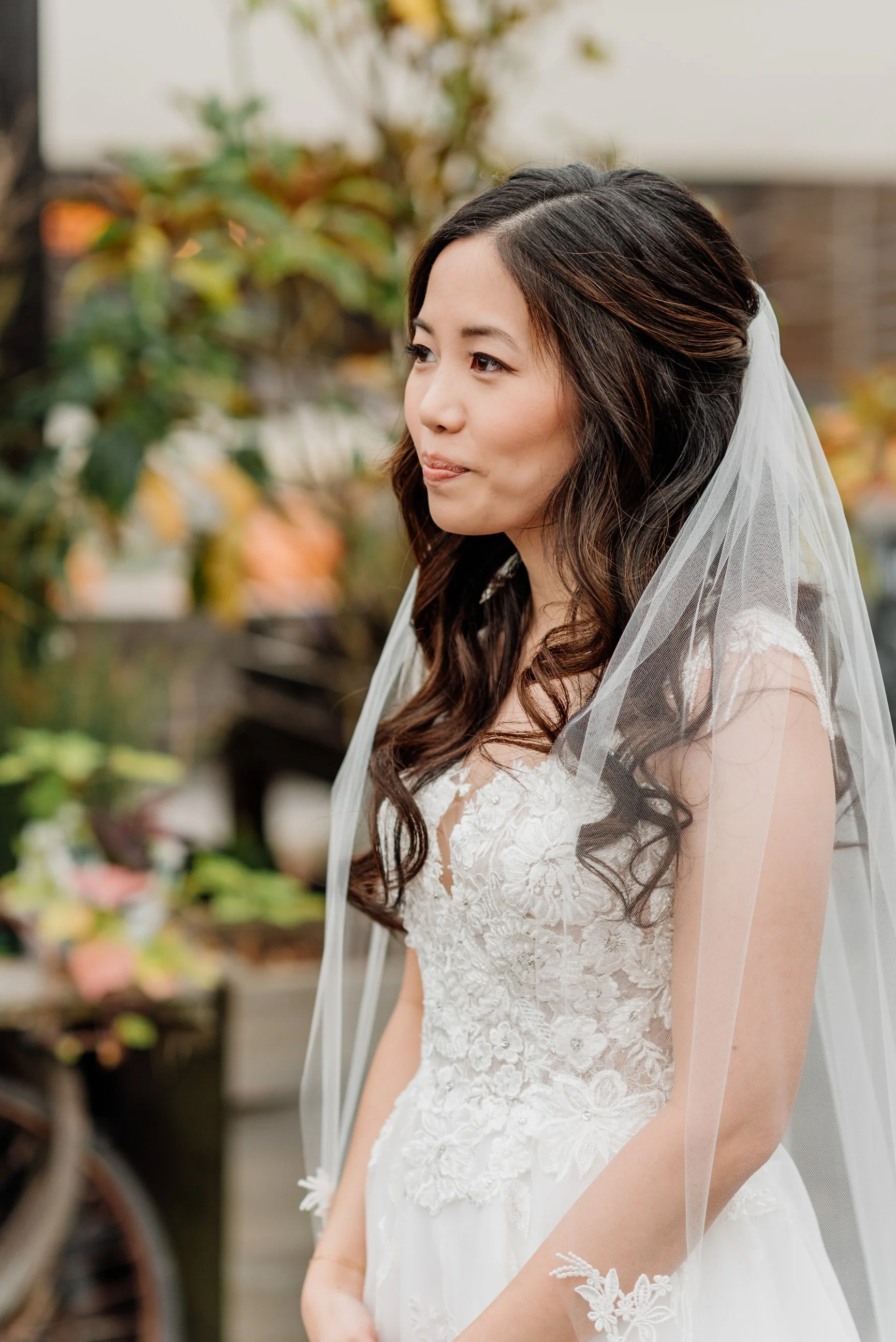 Bridal portrait of a woman with long dark wavy hair wearing a white lace wedding dress and a sheer veil, standing outdoors with blurred green and autumn-colored foliage in the background.