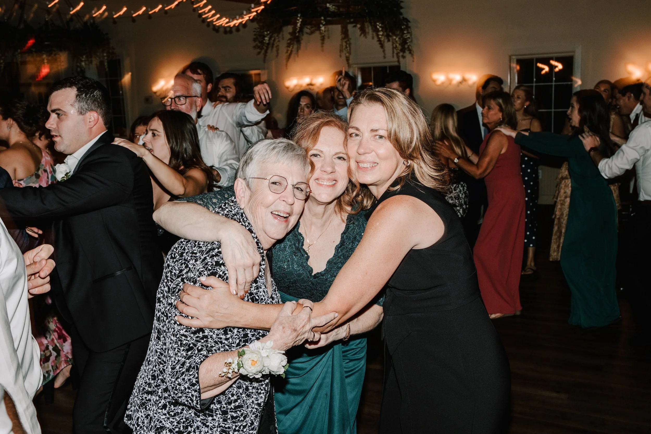 Three women hugging and smiling at a lively celebration or party, with many people dancing and enjoying themselves in the background.