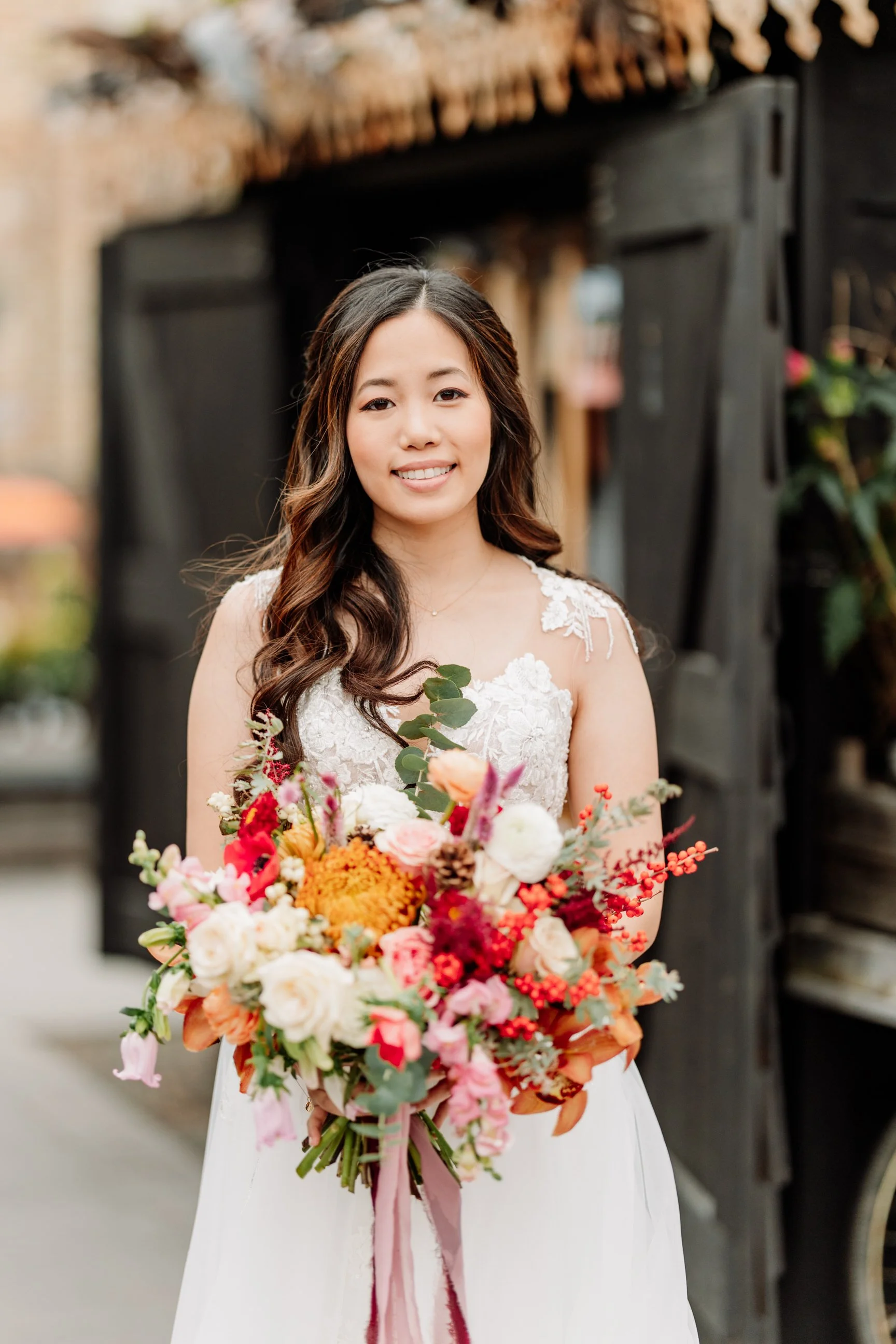 A woman in a wedding dress holding a colorful bouquet of flowers, standing outdoors near a rustic wooden structure.