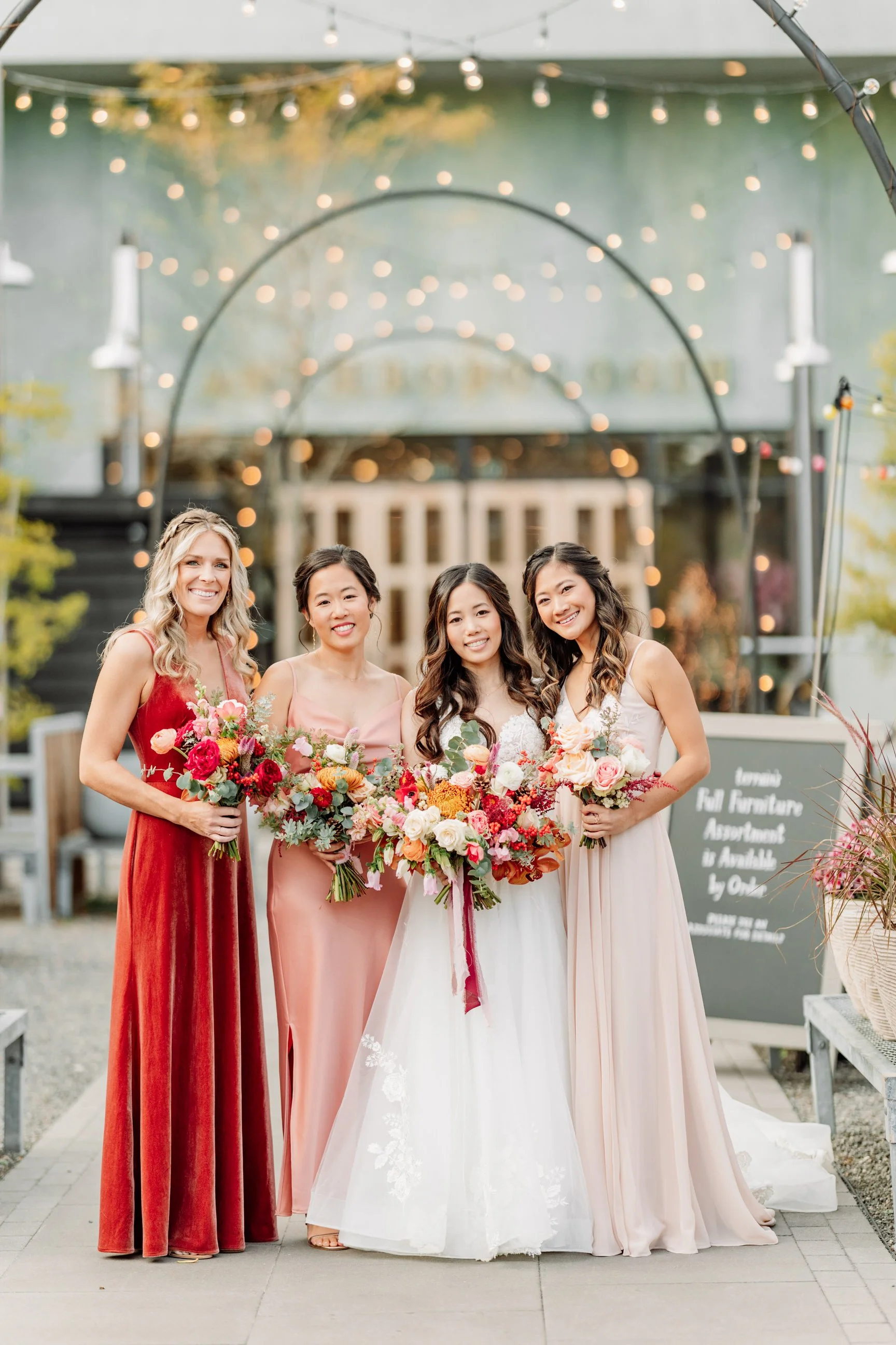 Four women in elegant dresses holding bouquets, standing outdoors at a wedding or celebration with string lights and decorative arches in the background.