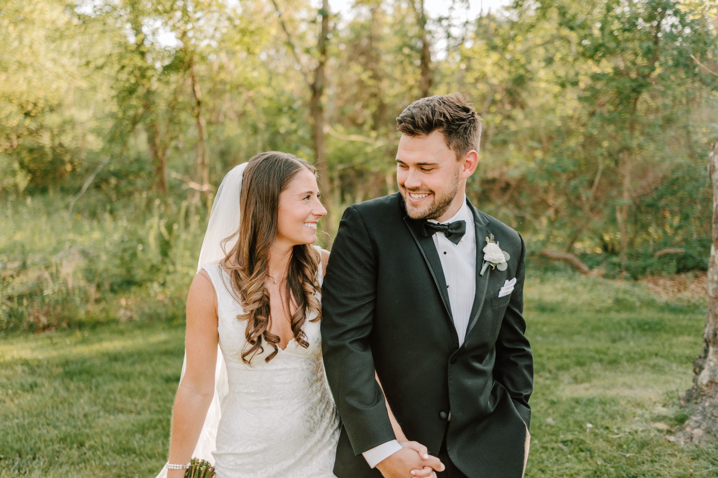 A bride and groom holding hands and smiling at each other outdoors in a park with green trees and grass, during their wedding celebration.