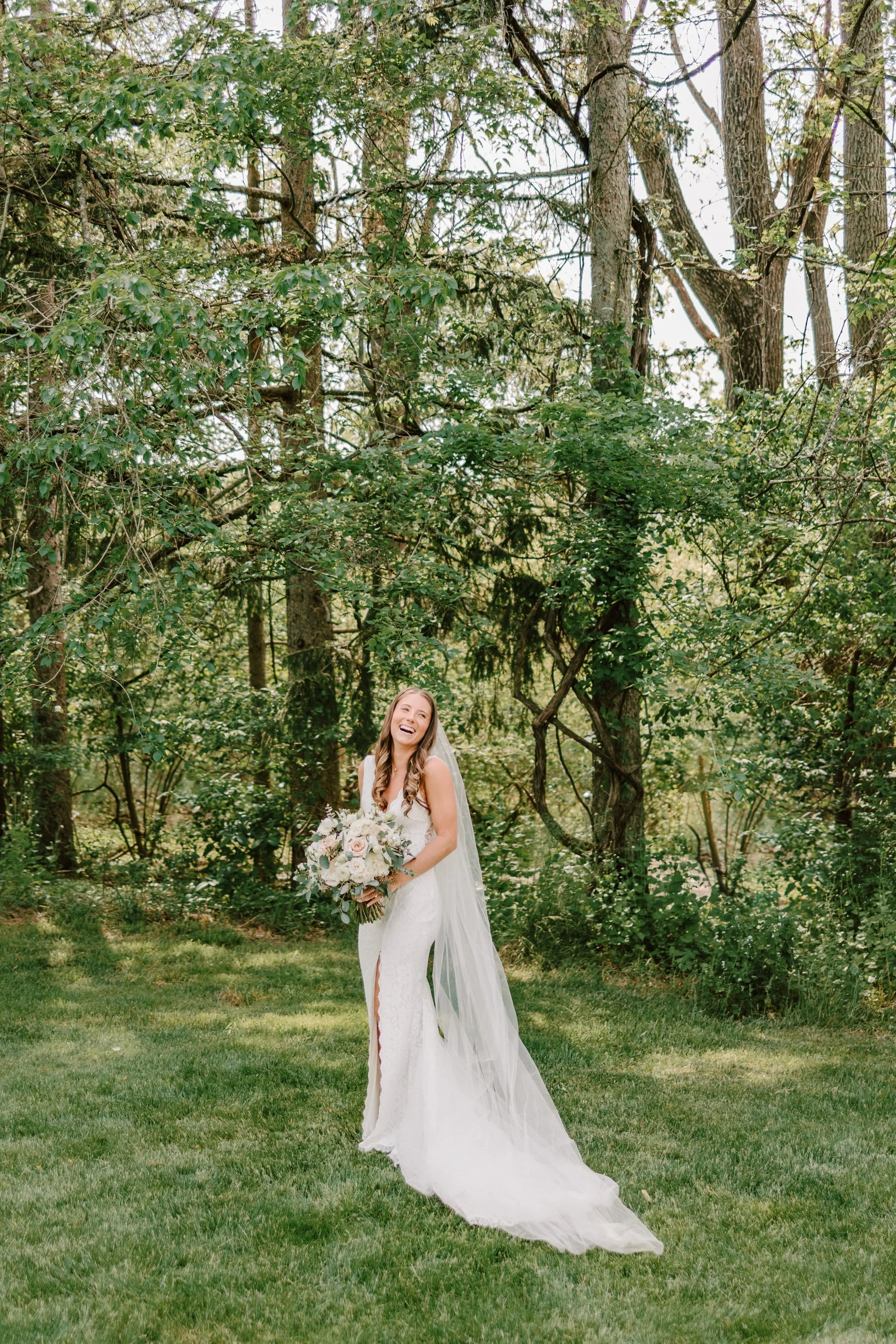 A bride in a white wedding dress holding a bouquet, standing on grass with trees in the background, smiling and laughing.
