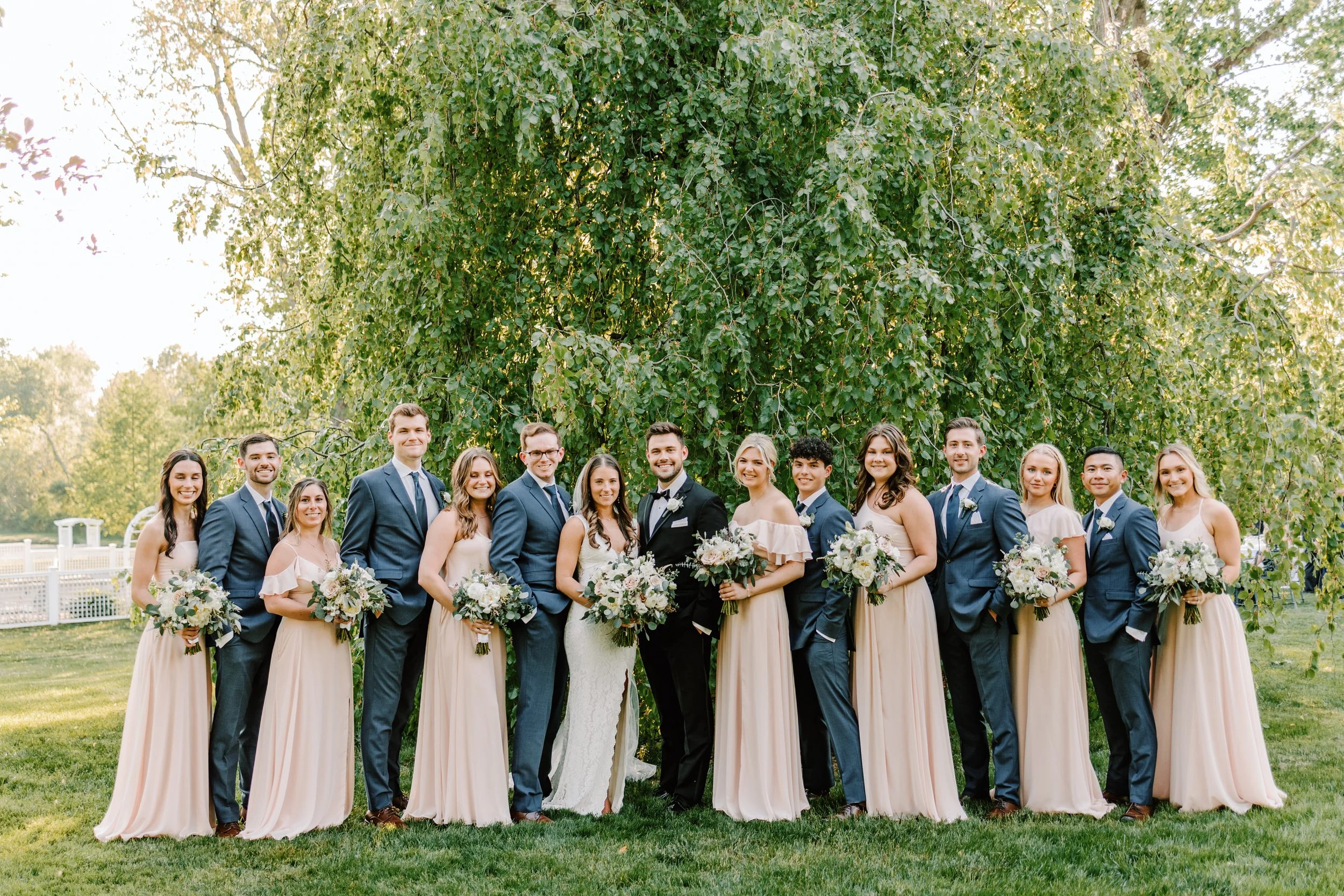 A wedding party standing outdoors under a large leafy tree, including the bride, groom, bridesmaids, and groomsmen holding bouquets and wearing formal attire. Philadelphia wedding photographer