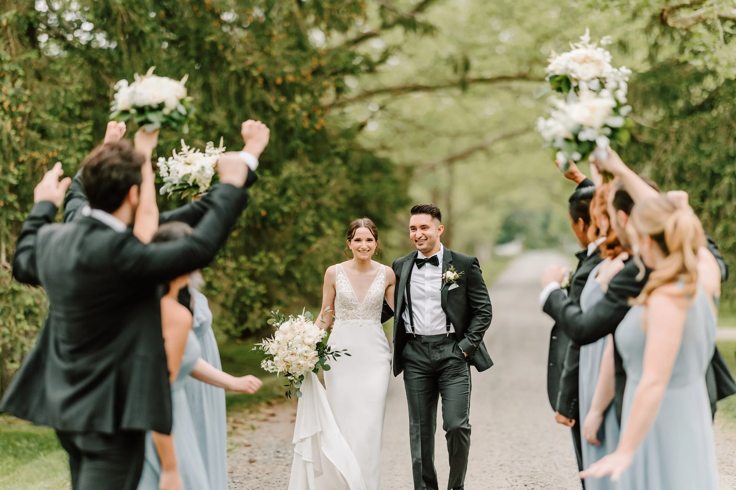 Bride and groom walking together outdoors, wedding party celebrating with bouquets and raised hands, trees in the background.