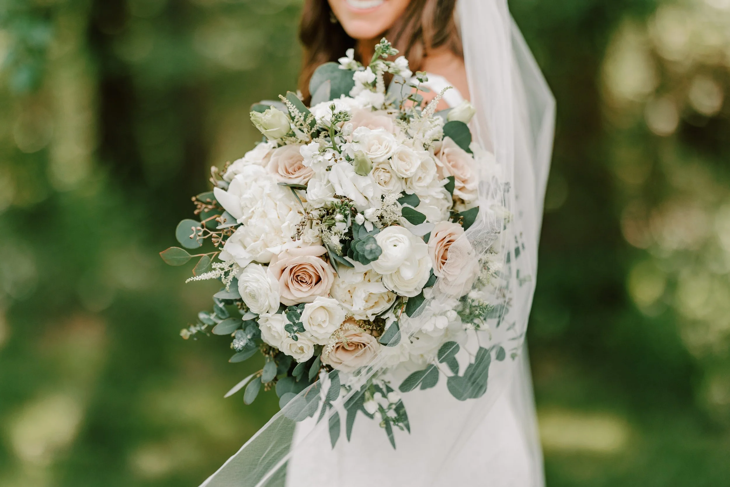 A bride holding a large bouquet of white and blush pink roses, ranunculus, and greenery, with a sheer veil, against a blurred outdoor background.