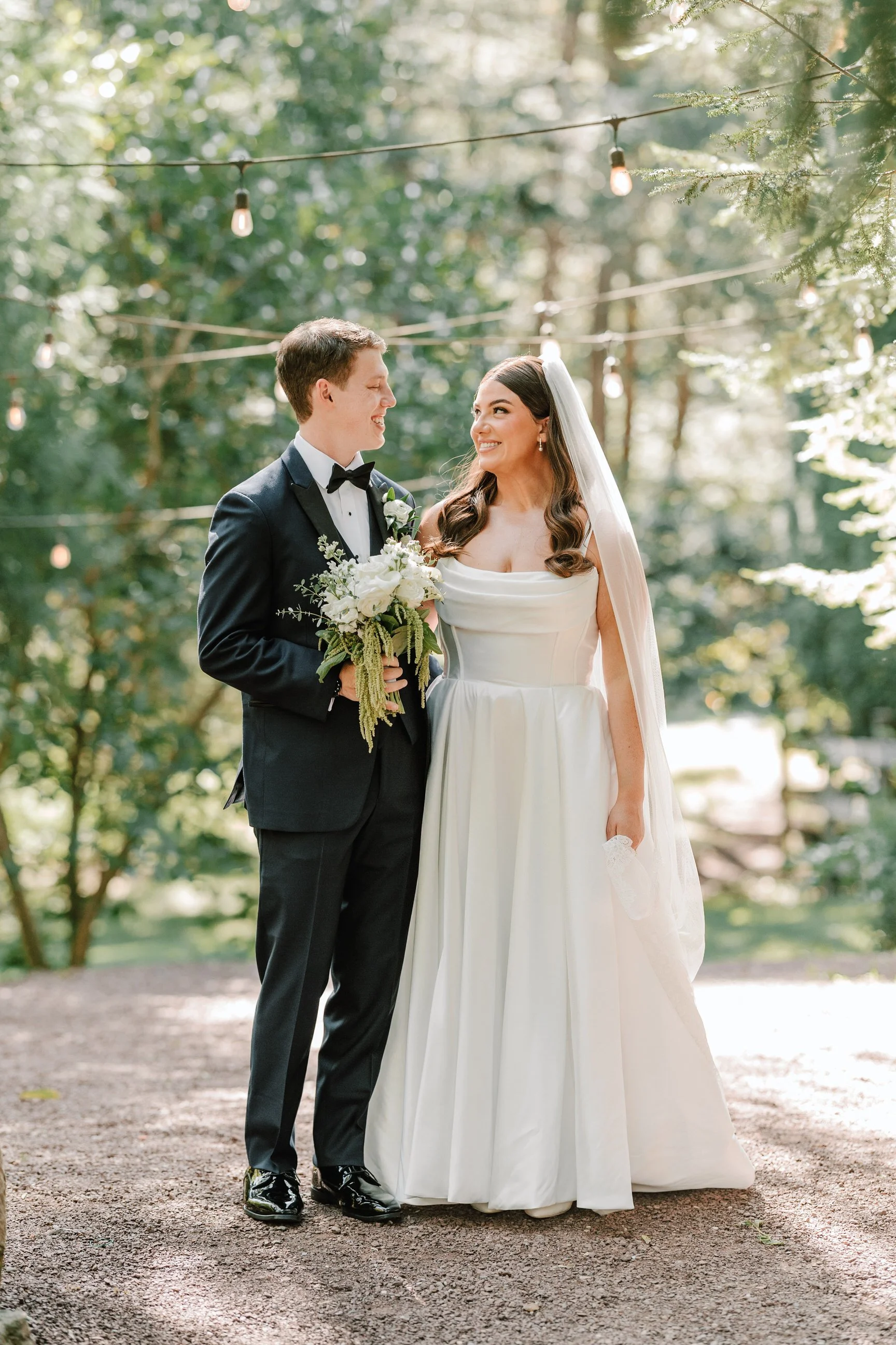 A bride and groom standing outdoors on their wedding day, surrounded by greenery, with string lights overhead. The groom is dressed in a black tuxedo holding a bouquet, and the bride is in a white wedding gown with a long veil, smiling at each other.