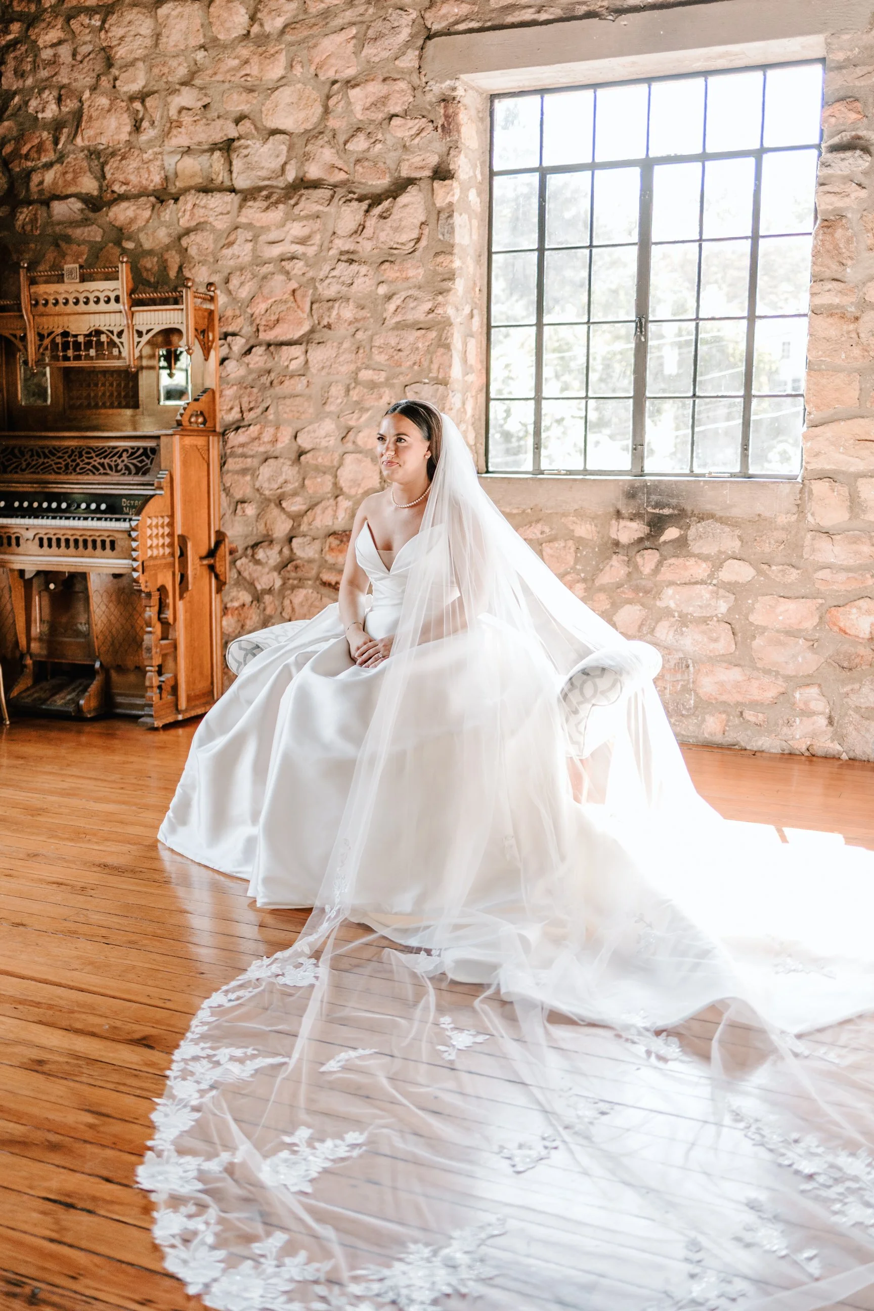 A bride sitting on a cushioned bench in a rustic room with stone walls and a large window, wearing a white wedding gown and veil.