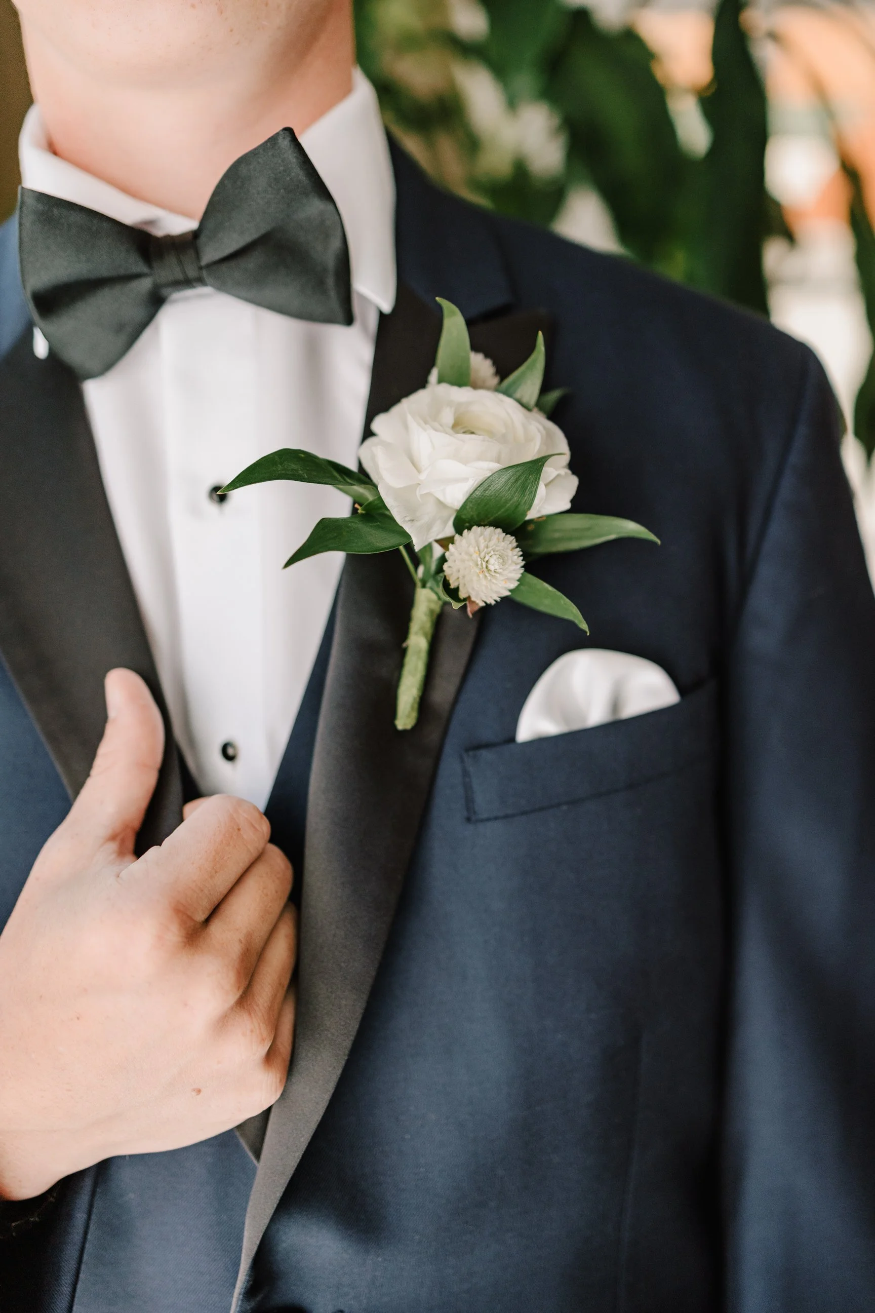 Close-up of a groom in a tuxedo with a black bow tie, white dress shirt, boutonniere with white flowers and green leaves, and a white pocket square.