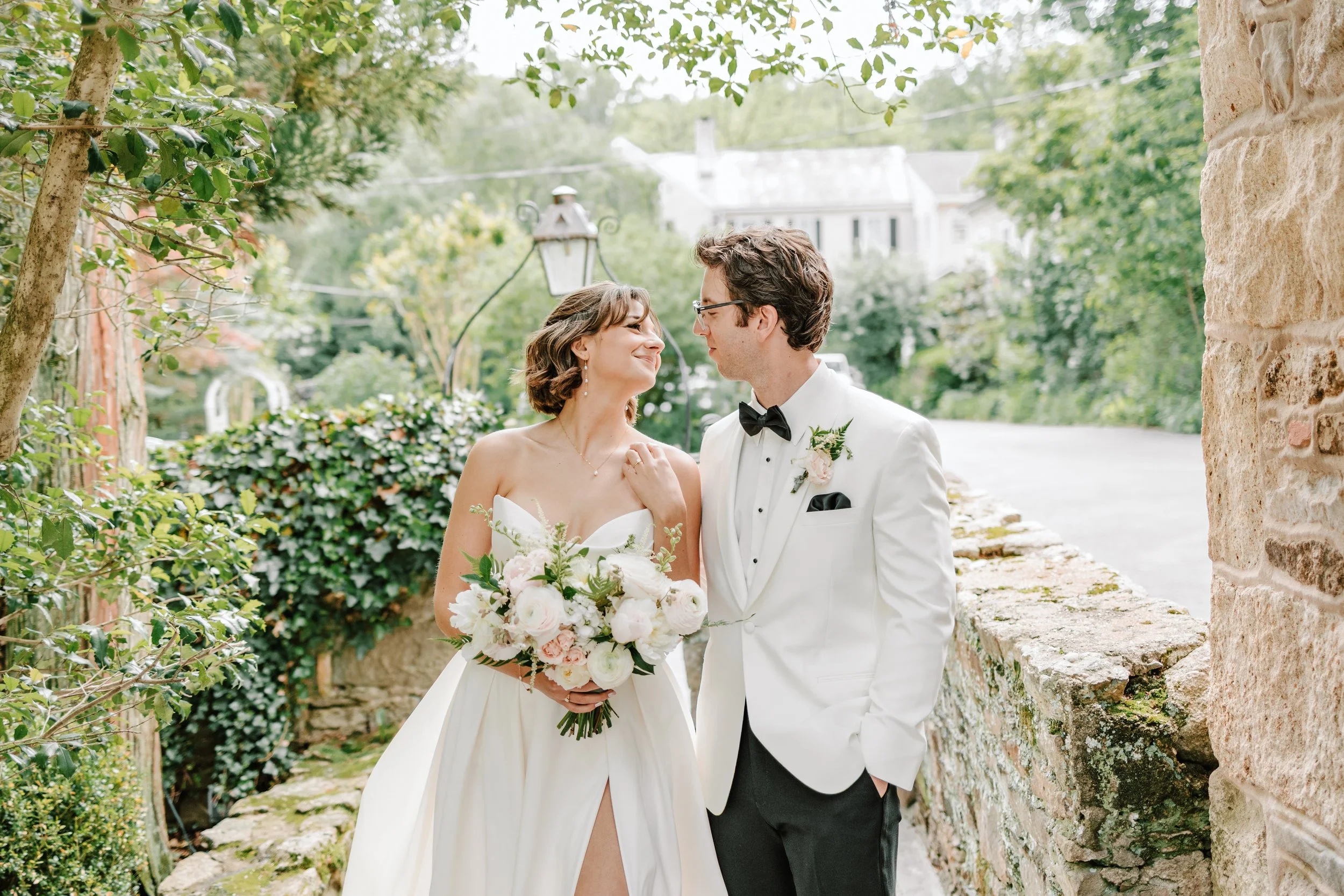 A bride and groom happily gazing at each other outdoors on their wedding day, with greenery and a stone wall in the background.