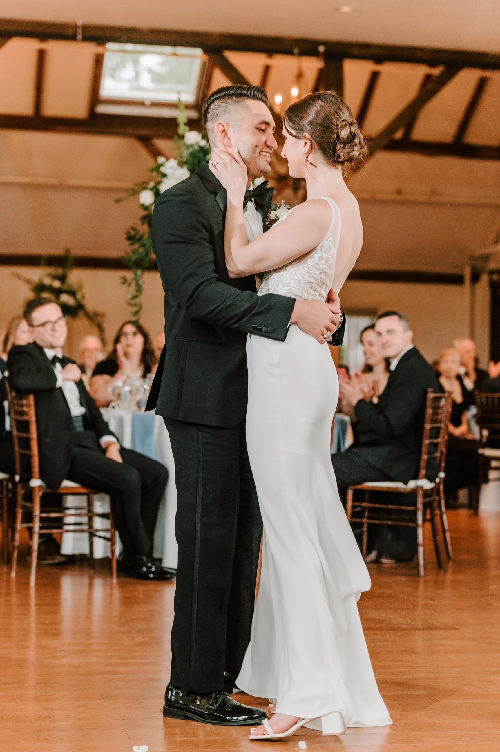 A bride and groom share their first dance at their wedding reception. The groom wears a black tuxedo, and the bride wears a white wedding gown with lace details. Guests are seated at tables, watching and clapping in the background.
