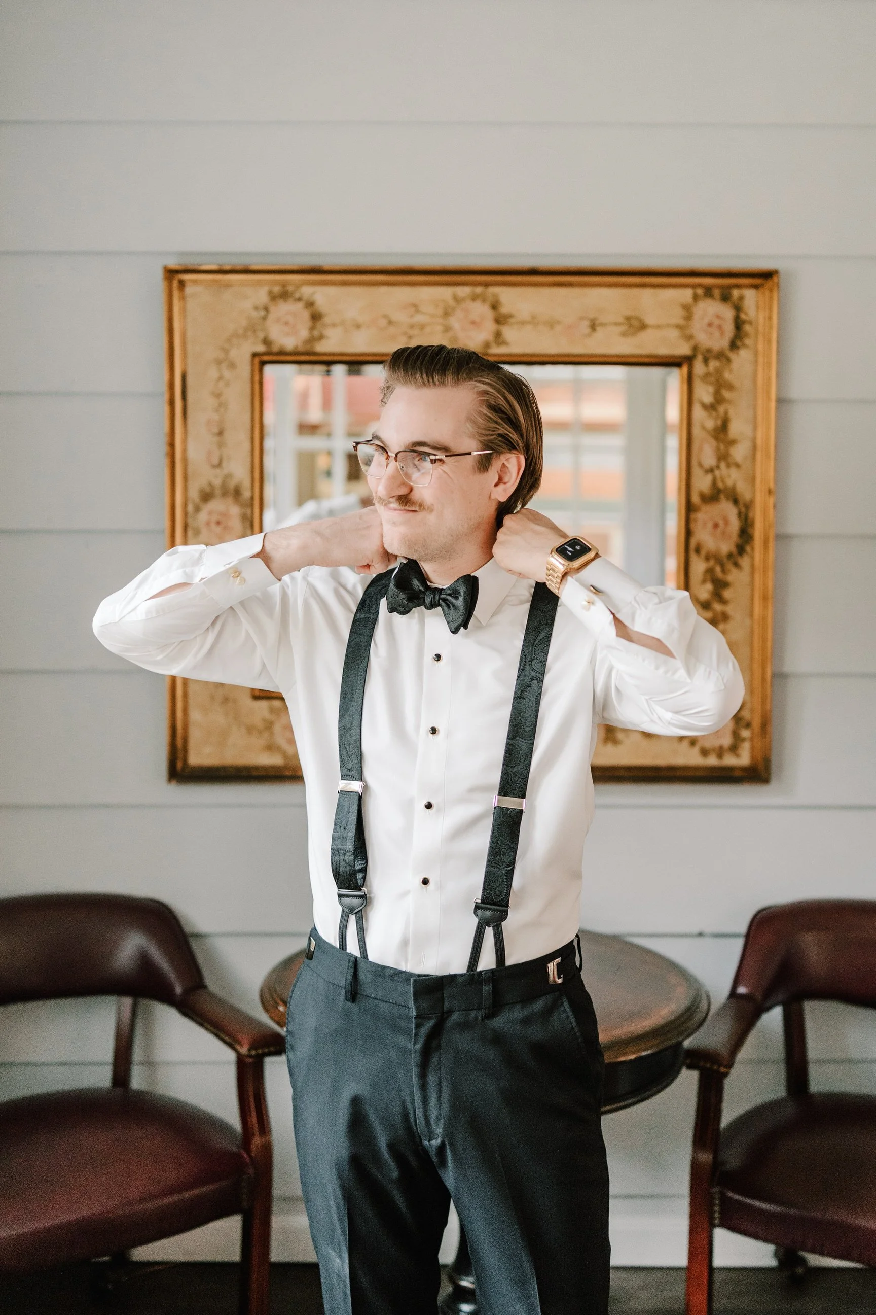 A man dressed in formal attire, adjusting his suspenders in front of a mirror. Holly Hedge Estate Sharyn Louise Photo