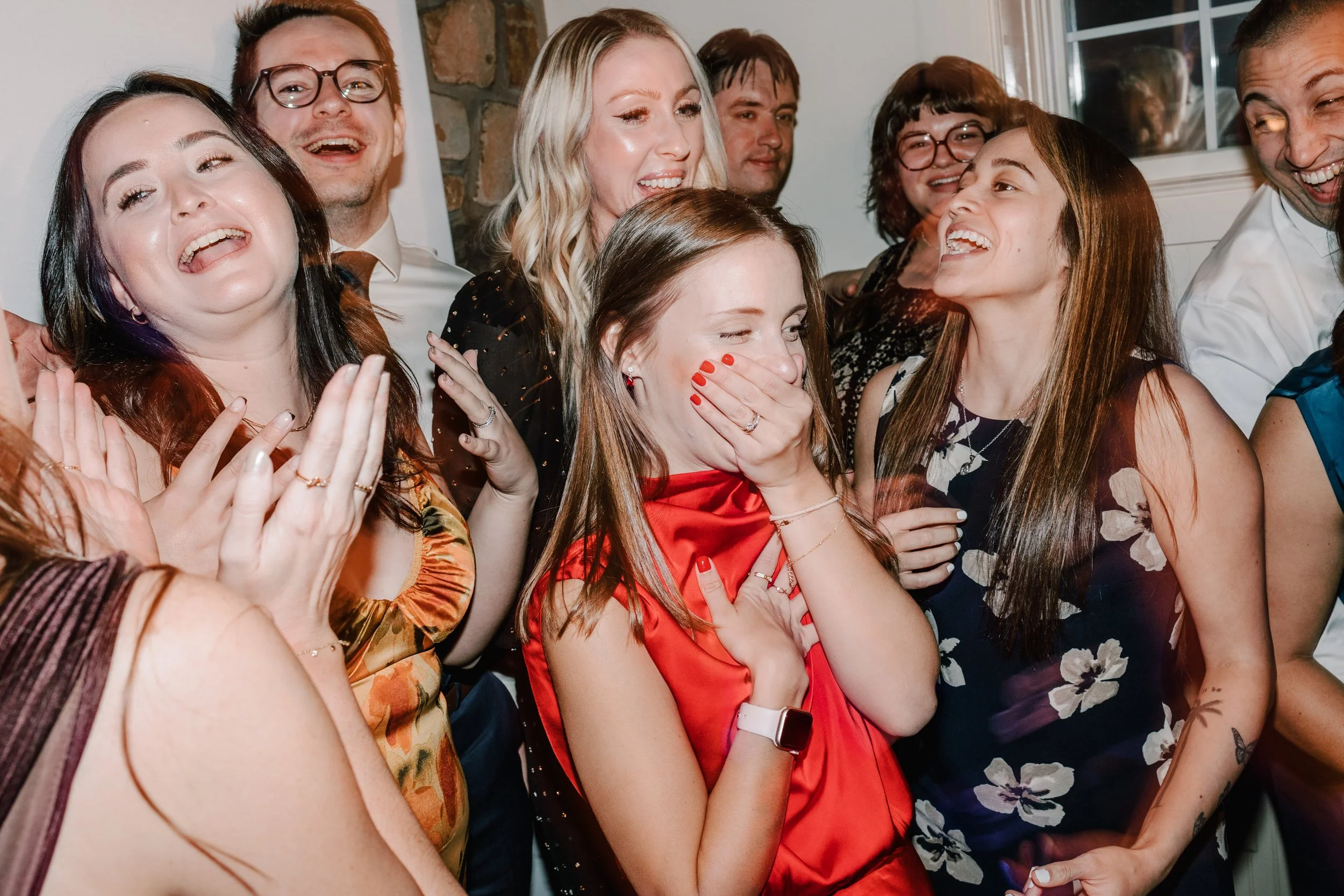 Group of people laughing and smiling at a celebration, with one woman covering her mouth in surprise.