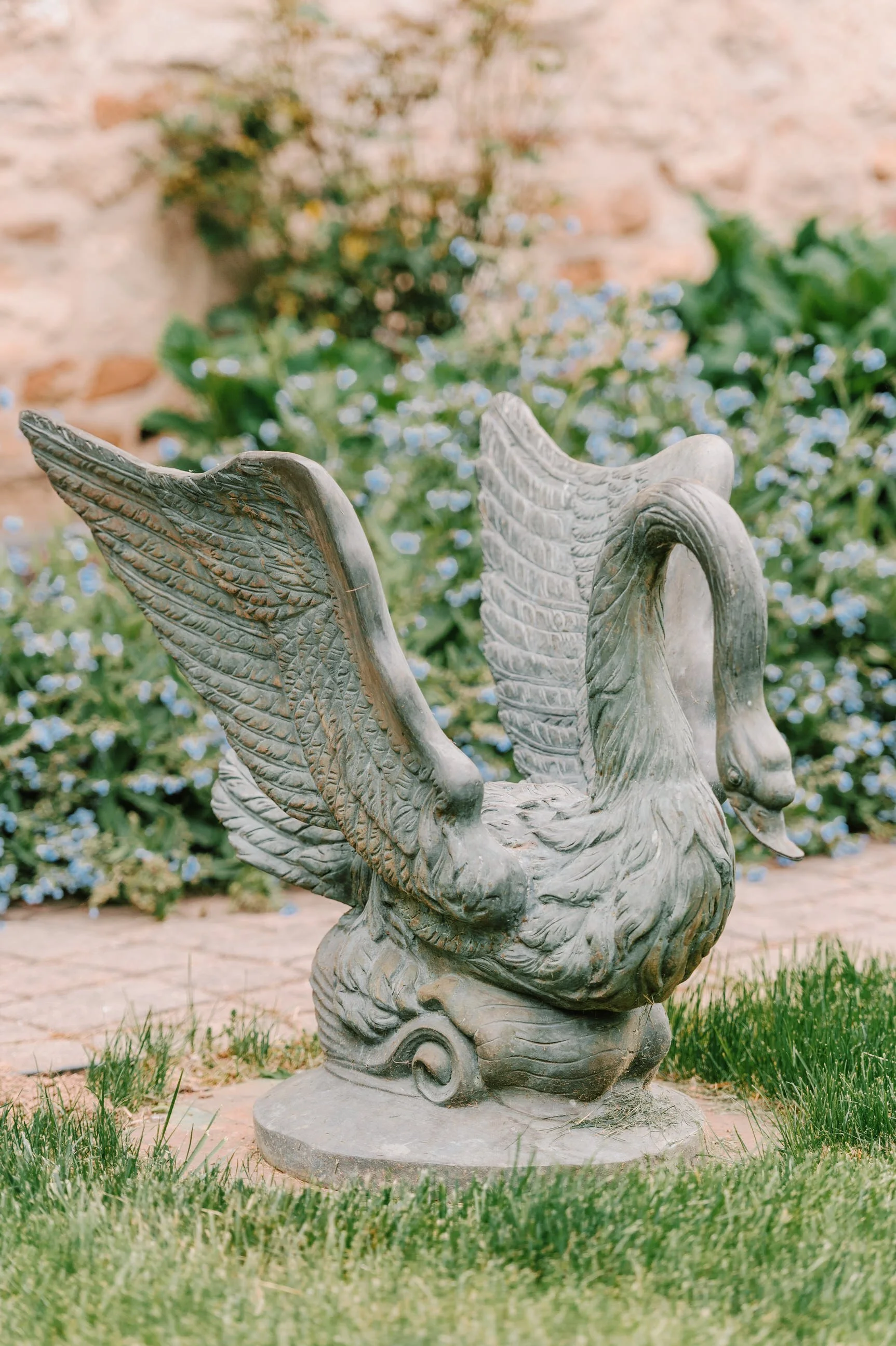A stone sculpture of a swan with detailed feathers in a garden with green grass and blooming flowers.