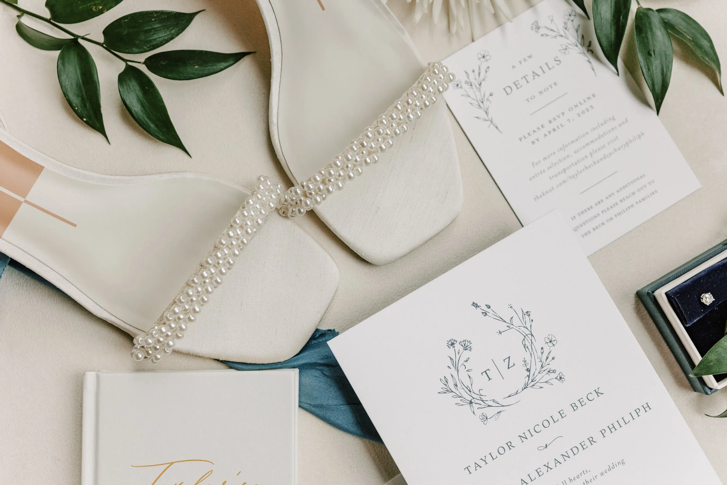 Wedding invitation suite, pearl headband, greenery, jewelry box, and decorative items arranged on a cream surface.