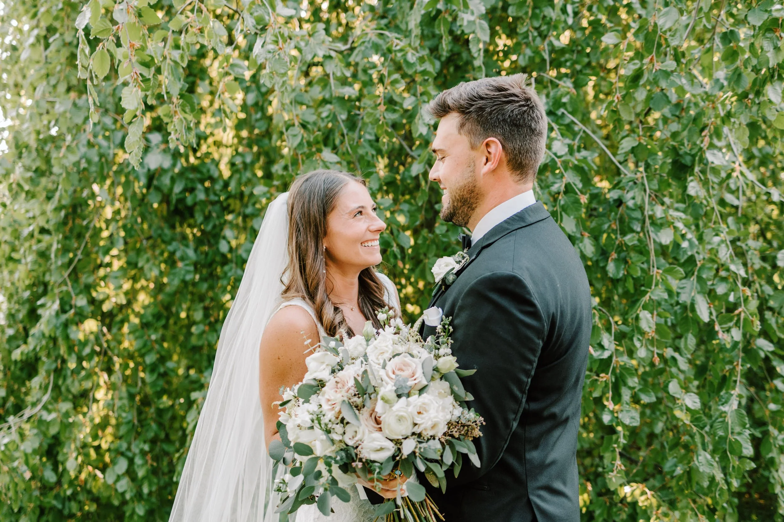 A bride and groom standing outdoors in front of green leafy trees, looking at each other and smiling. The bride is holding a large bouquet of white and blush flowers with greenery, wearing a veil. The groom is in a black tuxedo with a white boutonnie