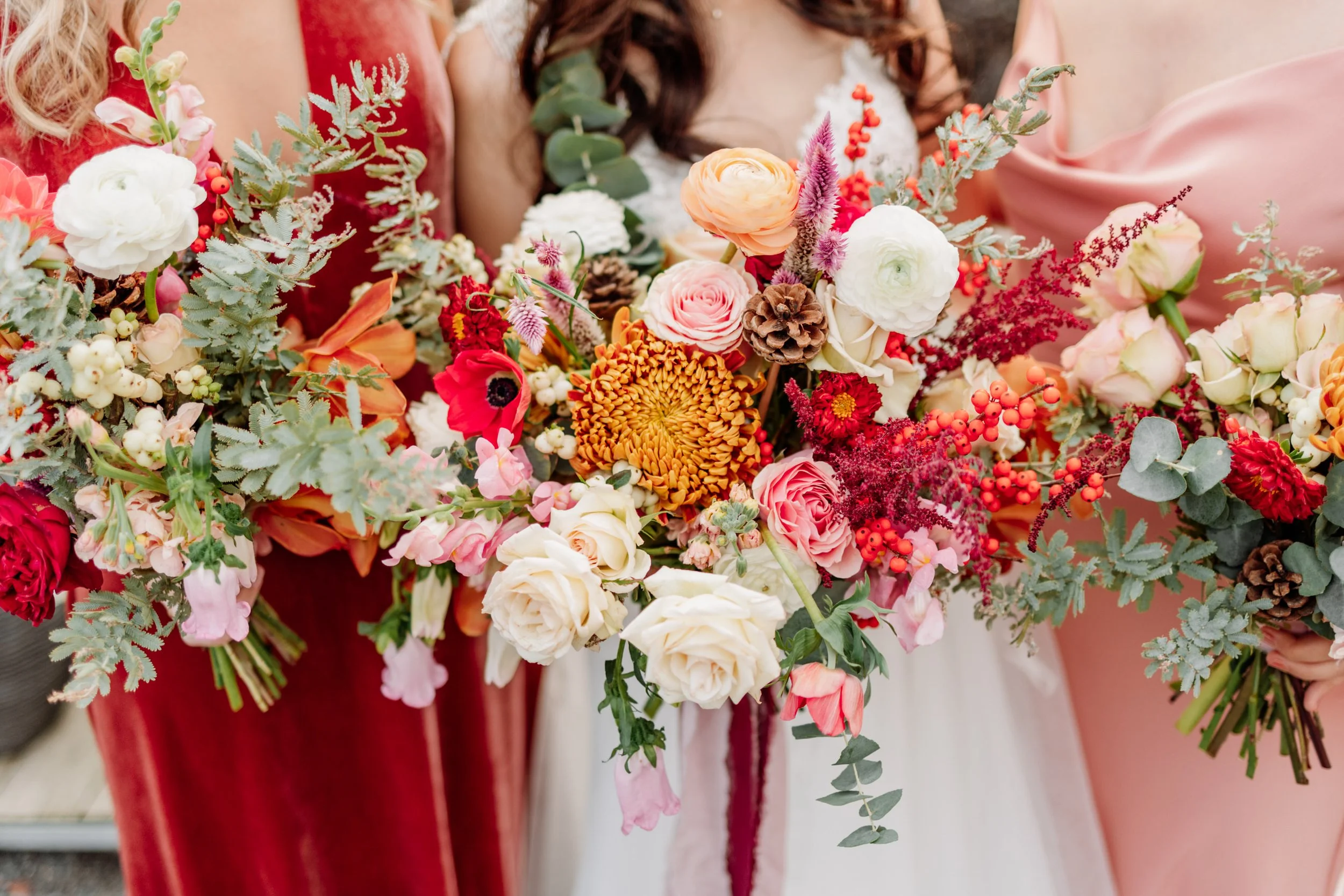 Close-up of two women holding colorful bouquets of flowers on an outdoor wedding day. The bouquets include roses, ranunculus, anemones, chrysanthemums, and assorted greenery.