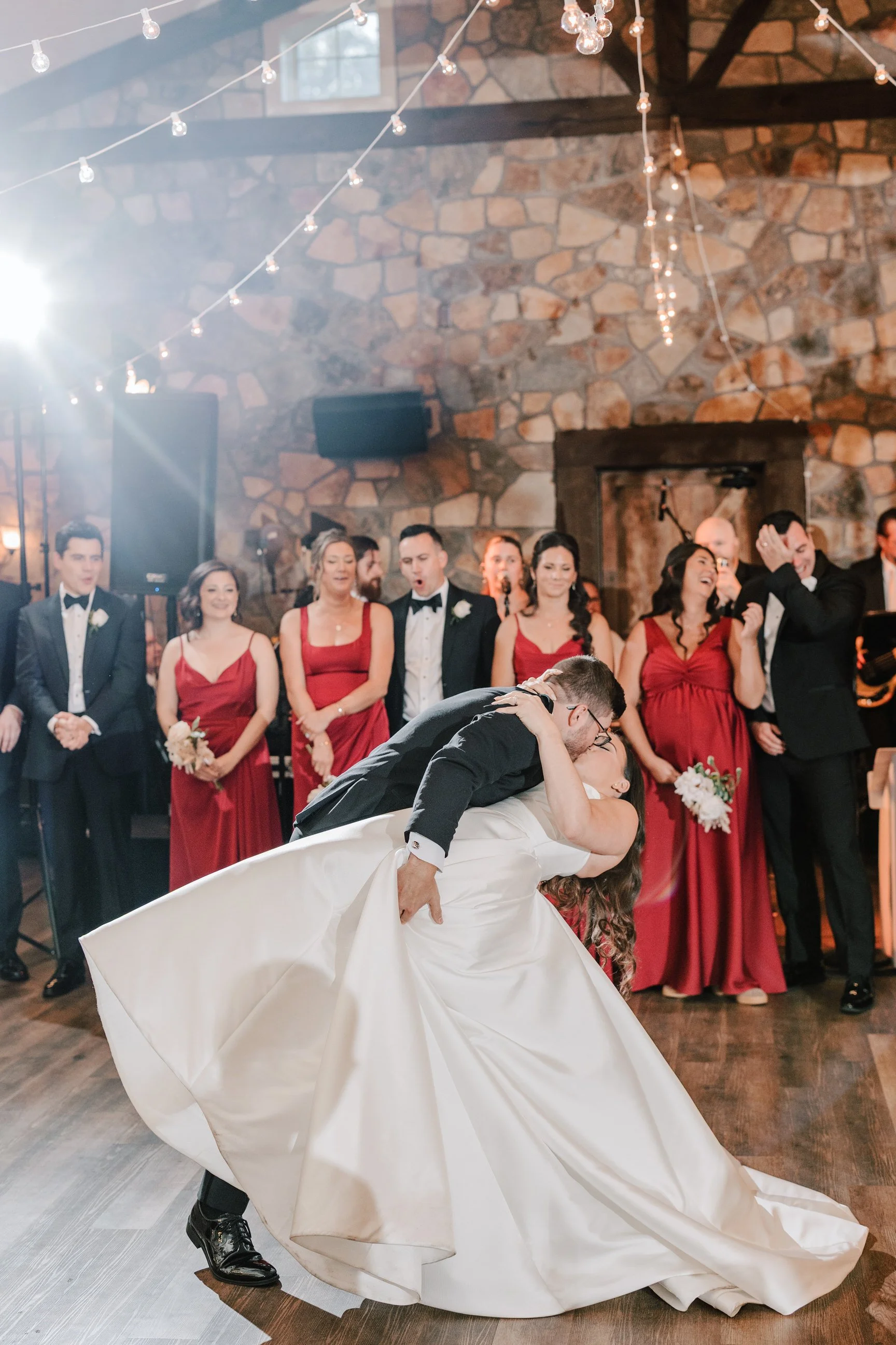 A bride and groom sharing a dip kiss during their wedding dance in a rustic venue with guests in formal attire watching and smiling.