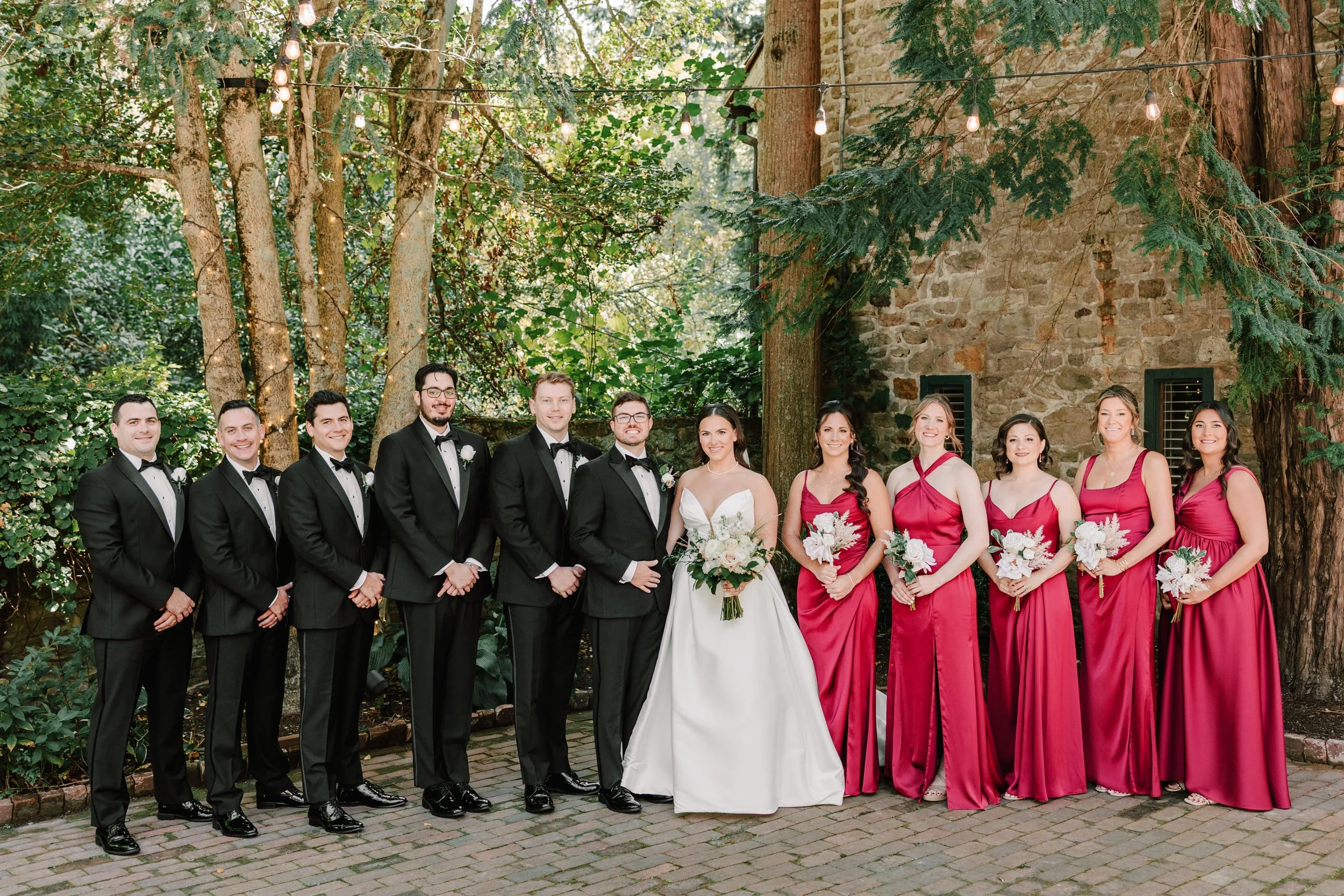 A wedding party standing outdoors, with nine men dressed in black tuxedos and ten women wearing pink bridesmaid dresses, posing on a brick patio surrounded by trees and a stone building.