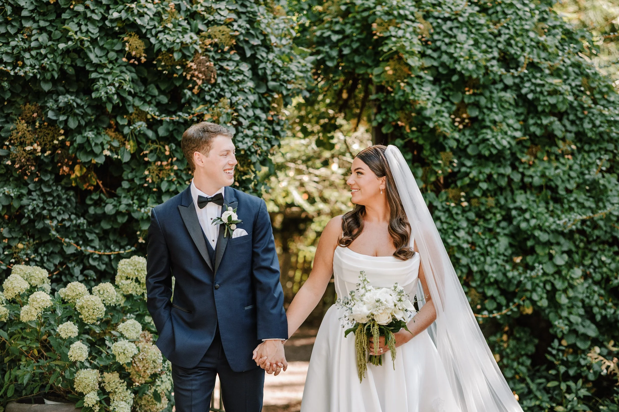 A newlywed couple holding hands and smiling at each other outdoors, with lush green foliage behind them. The groom is wearing a navy suit with a black bow tie and boutonniere, while the bride is in a white wedding dress with a long veil and holding a